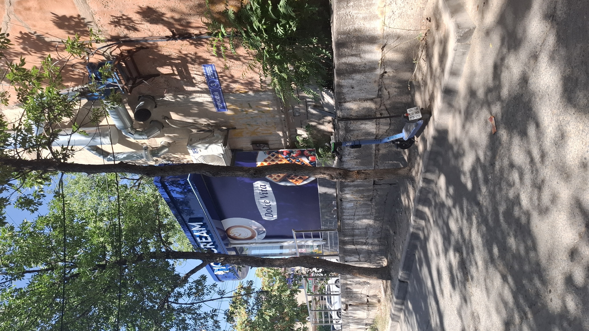 The image depicts an outdoor scene in the city of Shymkent, Kazakhstan, on a sunny day. In the foreground, a paved street is partially covered by dappled shadows cast by trees. A blue electric scooter with a license plate displaying "309 084" is parked on the sidewalk next to a concrete retaining wall. The retaining wall runs along the edge of a slightly elevated area.
Behind the wall, a blue storefront with "ICE CREAM" written in white lettering is visible. The storefront features a large purple mural with images of coffee and waffles. Two trees with green foliage stand in front of the storefront, partially obscuring it.
To the right of the storefront, the exterior wall of a building, colored in a reddish-orange hue, is present. This wall has exposed pipes and an air conditioning unit mounted on it. A street sign with Cyrillic lettering and the number "3" is affixed to the building wall. Green foliage grows around the base of the building. In the background, a white car is parked on a street beyond the retaining wall.