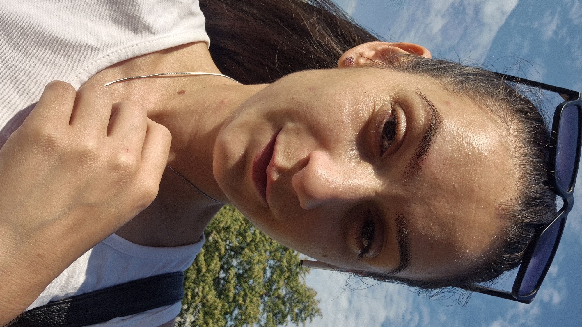 A selfie taken outdoors on a sunny day. The image shows a young woman with dark hair pulled back, wearing sunglasses perched on her head, and a white t-shirt. She has an earring in her left ear and is wearing a thin silver necklace. She has an earbud in her right ear. Her hand is raised and touching her neck. In the background, there is a blue sky with white clouds and a green tree. There are no visible signs of the location being in the city of Sofia, Bulgaria, or any specific urban setting. The focus is on the woman's face and upper body.