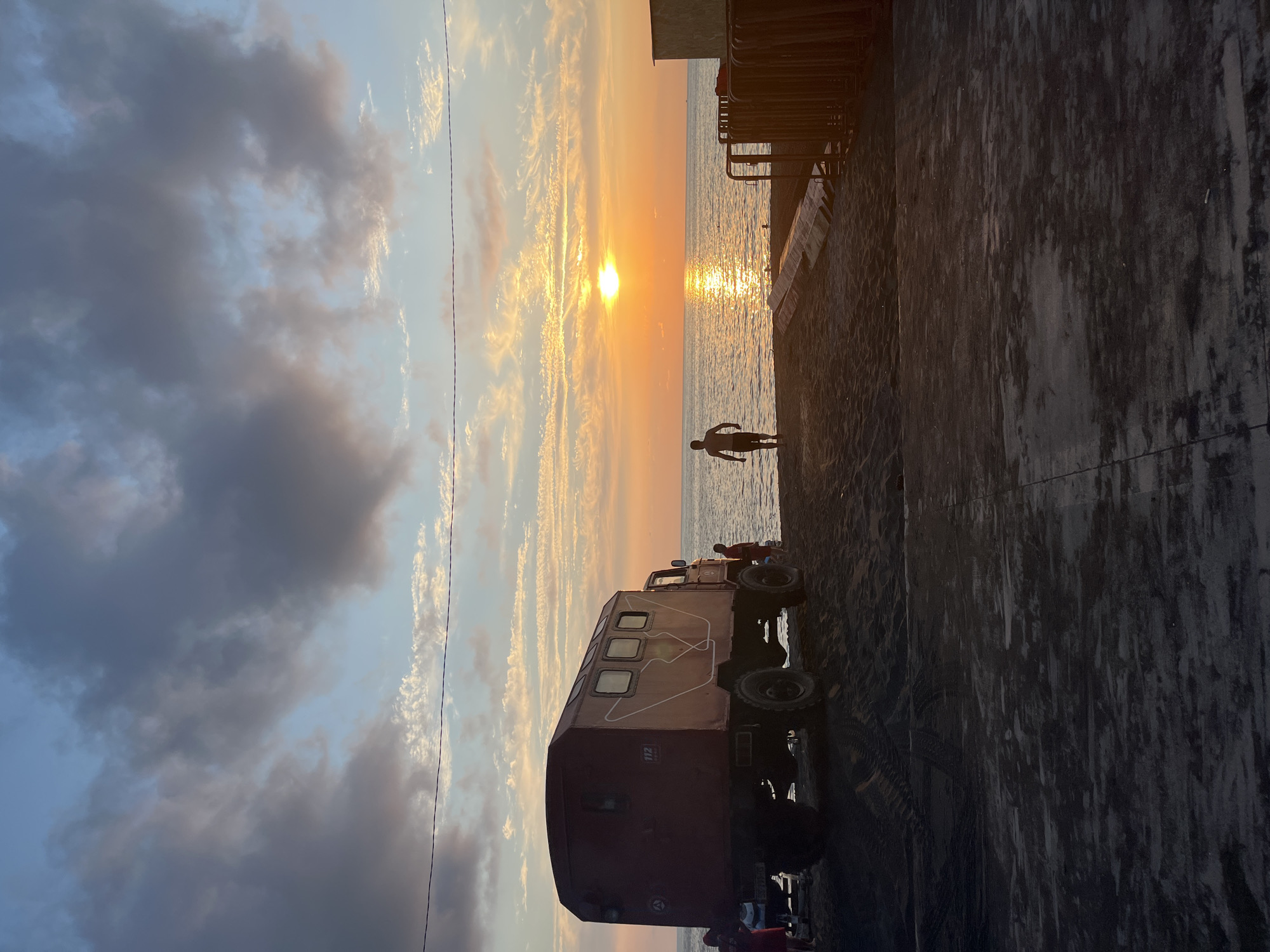 A sunset scene on the beach in Ureki, Georgia, features a large, maroon utility truck parked on the dark sand. The truck has several windows and a number "112" painted on its side. To the left of the truck, two figures, one wearing red, are partially visible. A lone man, silhouetted against the setting sun, walks towards the water. The ocean stretches out to the horizon, reflecting the vibrant orange and yellow hues of the sunset. The sky is a blend of blue and grey clouds, with streaks of white and pink. In the foreground, dark sand is marked with tire tracks, and a ramp-like structure leads towards the water. To the right, a section of a building or structure and a row of metal poles are visible.