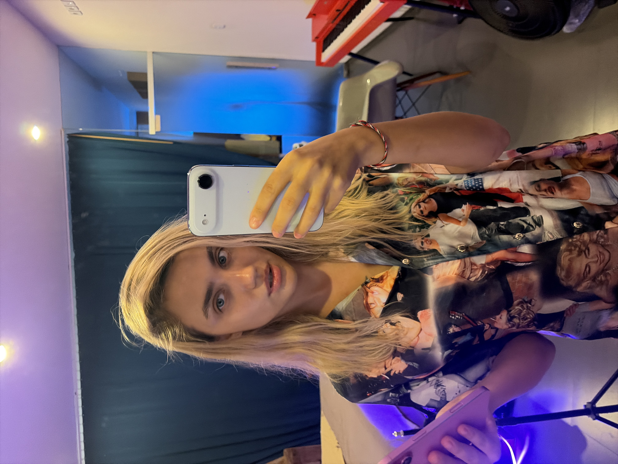 A young woman with blonde hair is taking a selfie in what appears to be a studio or dressing room. She is wearing a short-sleeved, collared shirt with a collage print of various people. She is holding a white smartphone in her right hand, capturing her reflection in a mirror. In her left hand, she holds a pink object, possibly a phone case or tablet. The background shows a dark teal curtain, a chair, a red keyboard, and a blue-lit wall. The lighting is a mix of warm overhead light and cool blue light, creating a dramatic atmosphere. The location is not specified as Mengwi, Indonesia.