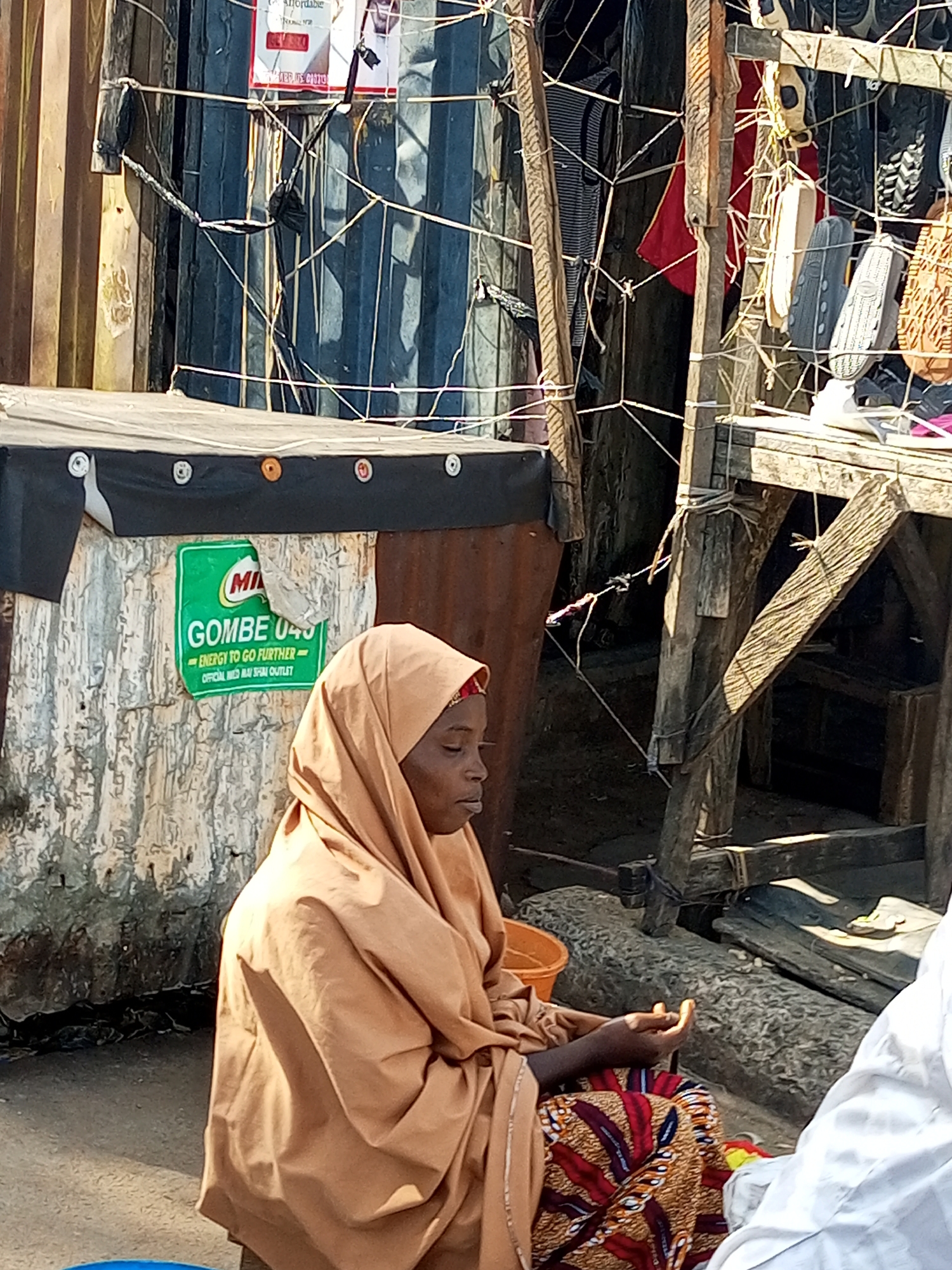 A woman wearing a light brown hijab and a colorful patterned skirt sits on the ground in front of a corrugated metal wall. She is looking down with her hands clasped. To her left, a green sign with the Milo logo and text reads "GOMBE 045 ENERGY TO GO FURTHER OFFICIAL MED AY SHEM OUTLET". Behind her, there is a wooden structure with various items, including what appear to be shoes or sandals, hanging from it. The environment appears to be an outdoor market or street stall in Gombe, Nigeria.