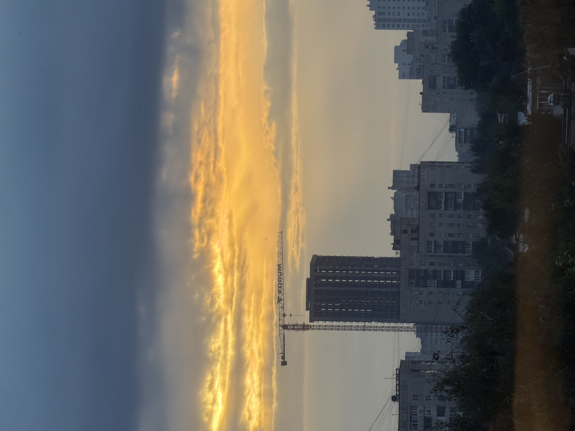The image depicts a view of a construction site with a crane and unfinished buildings against a dramatic sky. The sky is divided into two sections: a dark, ominous gray cloud layer at the top, and a vibrant, golden-orange band of sunlight breaking through below, illuminating scattered clouds. In the middle ground, a prominent red and yellow construction crane with the text "FORUM" is positioned over a tall, dark, unfinished building that appears to be a high-rise apartment complex under construction. To the left and right of this central structure are other residential buildings, some appearing complete and others also under construction or renovation. These buildings are typical of Soviet-era apartment blocks, characterized by their concrete facades and repetitive window patterns. In the foreground, a dense area of dark green foliage, likely trees and bushes, obscures the lower parts of the buildings. Beyond the foliage, a blurred band of dry grass or reeds is visible, suggesting a natural or park-like area in front of the urban landscape. The overall impression is one of urban development taking place amidst a striking natural phenomenon of light and cloud. While the image is not geographically explicit, the construction company name "FORUM" may refer to a development company operating in Russia. Without further contextual information or identifying landmarks, it is not possible to definitively identify the specific location within Yekaterinburg, Russia.