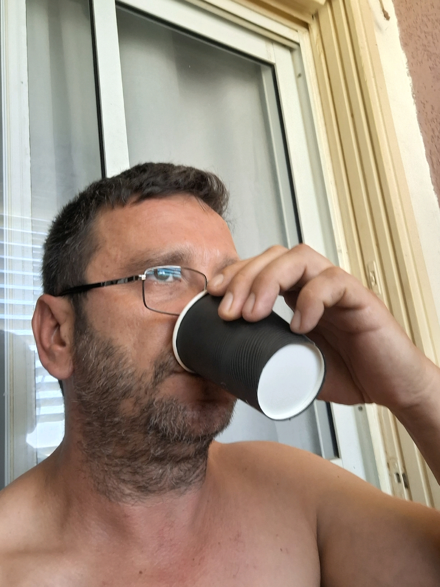 A man with short, dark hair and a beard, wearing glasses, is holding a black paper cup to his lips and drinking from it. He is outdoors, with a window and a light-colored wall in the background. The image appears to be taken in Yerocham, Israel.