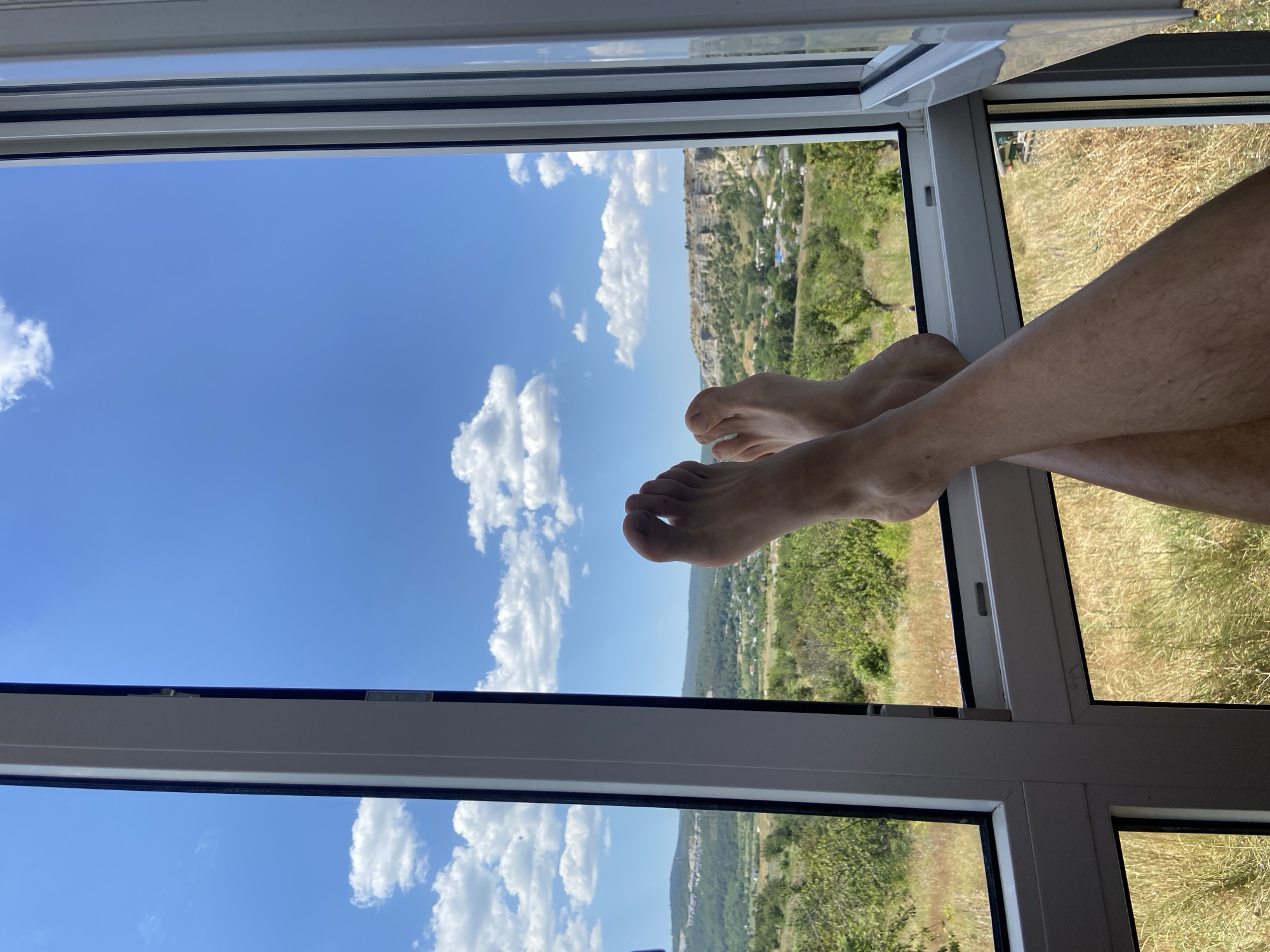 A first-person perspective shot shows a person's legs and feet resting with their ankles crossed, obscuring the lower portion of the frame. The feet are bare and positioned in front of a window that offers a view of the Krasnomakskoye rural settlement in Ukraine. The landscape beyond the window is characterized by rolling hills covered in green vegetation and scattered trees. A village with buildings is visible nestled in the valley. The sky is a vibrant blue, punctuated by fluffy white clouds. The window frame, made of white material, divides the scene.
