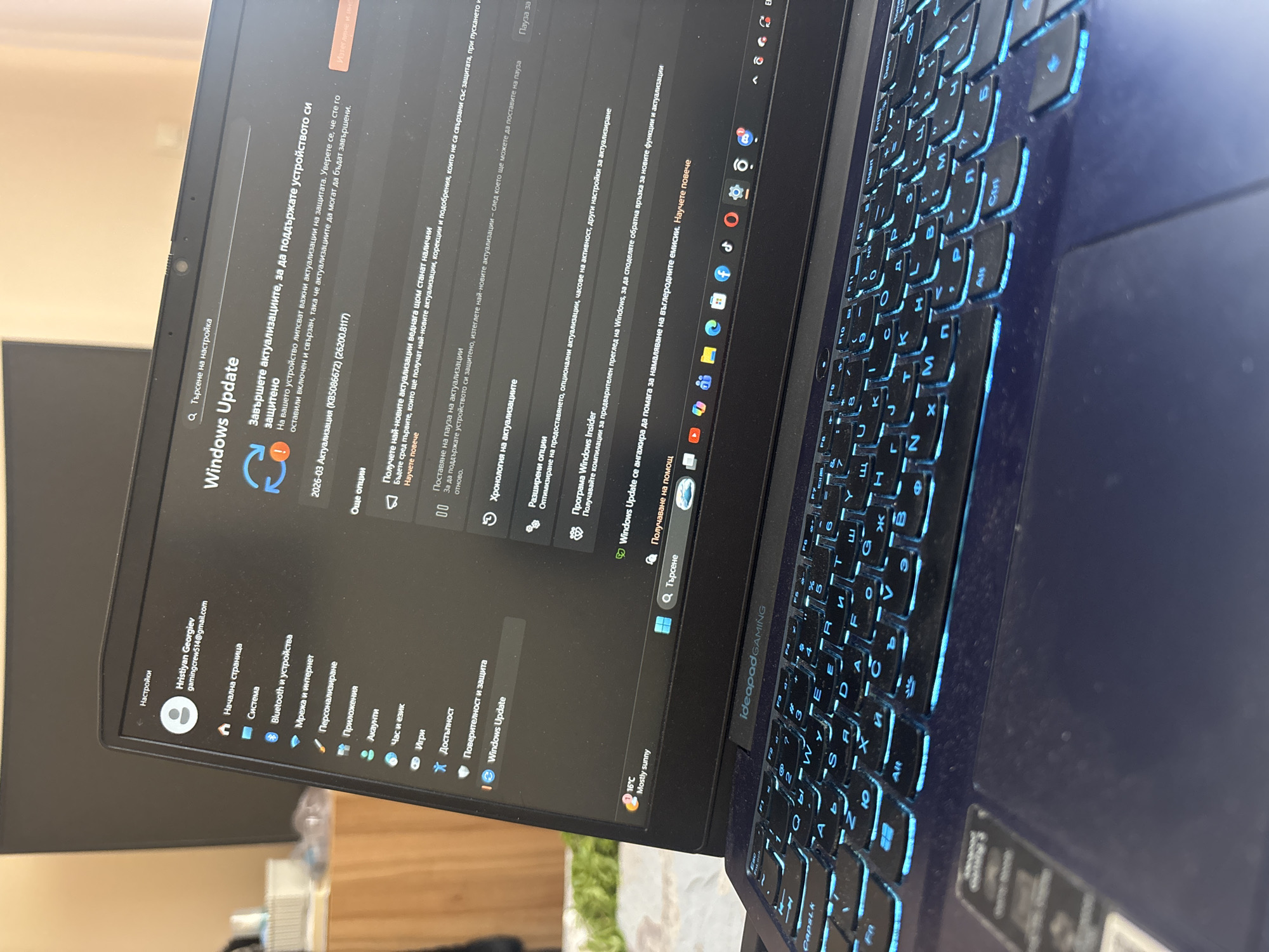 The image shows a close-up view of a laptop, possibly a Lenovo Ideapad Gaming model, displaying the Windows Update settings screen in Bulgarian. The screen indicates that updates are required to maintain the device's security. The keyboard is illuminated with blue backlighting, and the touchpad is visible. The laptop is open, and the screen is at an angle, making the bottom part of the keyboard visible. The system tray shows a temperature reading of 16°C and a "mostly sunny" weather condition. The laptop is on a surface, with a glimpse of an ambiguous object in the background.