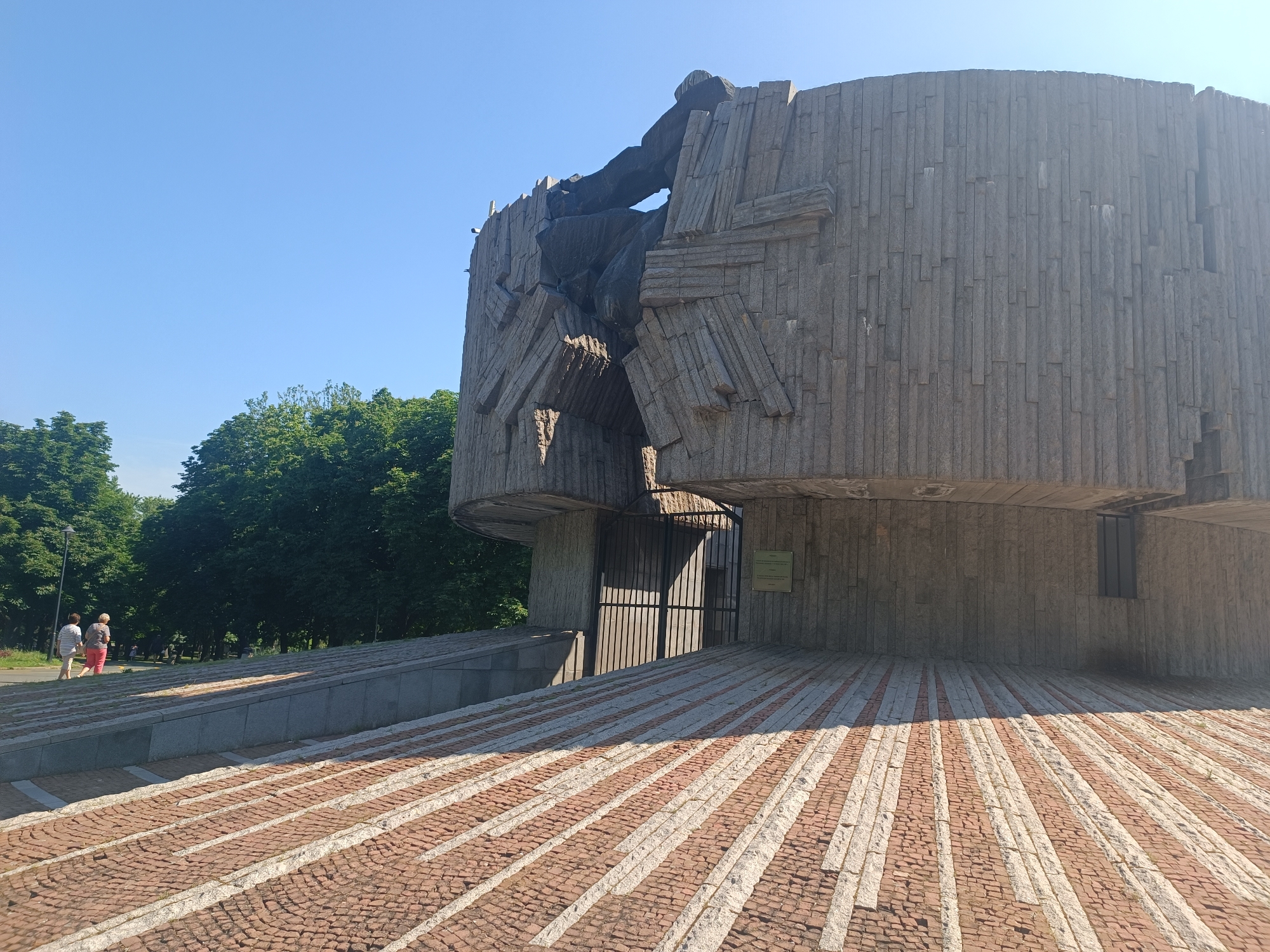 The image shows the Pantheon of the National Revival heroes in Burgas, Bulgaria. It is a monumental structure made of concrete and stone, with a unique, abstract design featuring a large opening at the top that appears to be breaking apart. The building is surrounded by trees and a clear blue sky. In the foreground, there is a patterned walkway made of red and white stones. In the background, two people are walking on a path. A metal gate leads into the building. A sign is mounted on the wall next to the gate. The overall impression is one of a solemn and impressive monument.