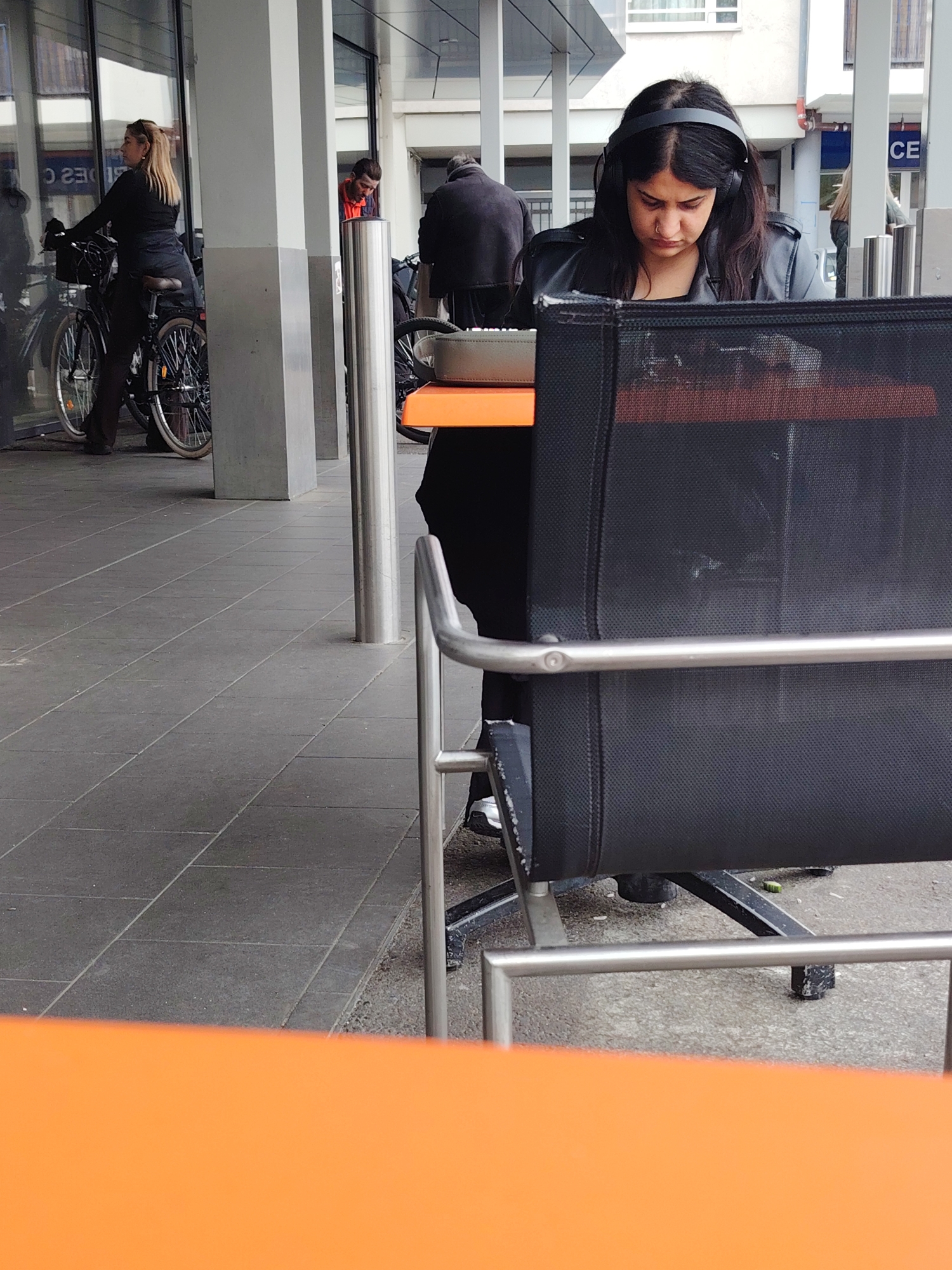 The image captures a scene in Strasbourg, France. A woman with dark hair, wearing headphones and a dark jacket, sits at an outdoor table with an orange top. She is partially obscured by a black mesh chair, looking down at something on the table. In the background, there are other pedestrians, some near bicycles parked by a building with glass windows. The ground is paved with gray tiles. A building is visible in the background with a sign that reads "CE."