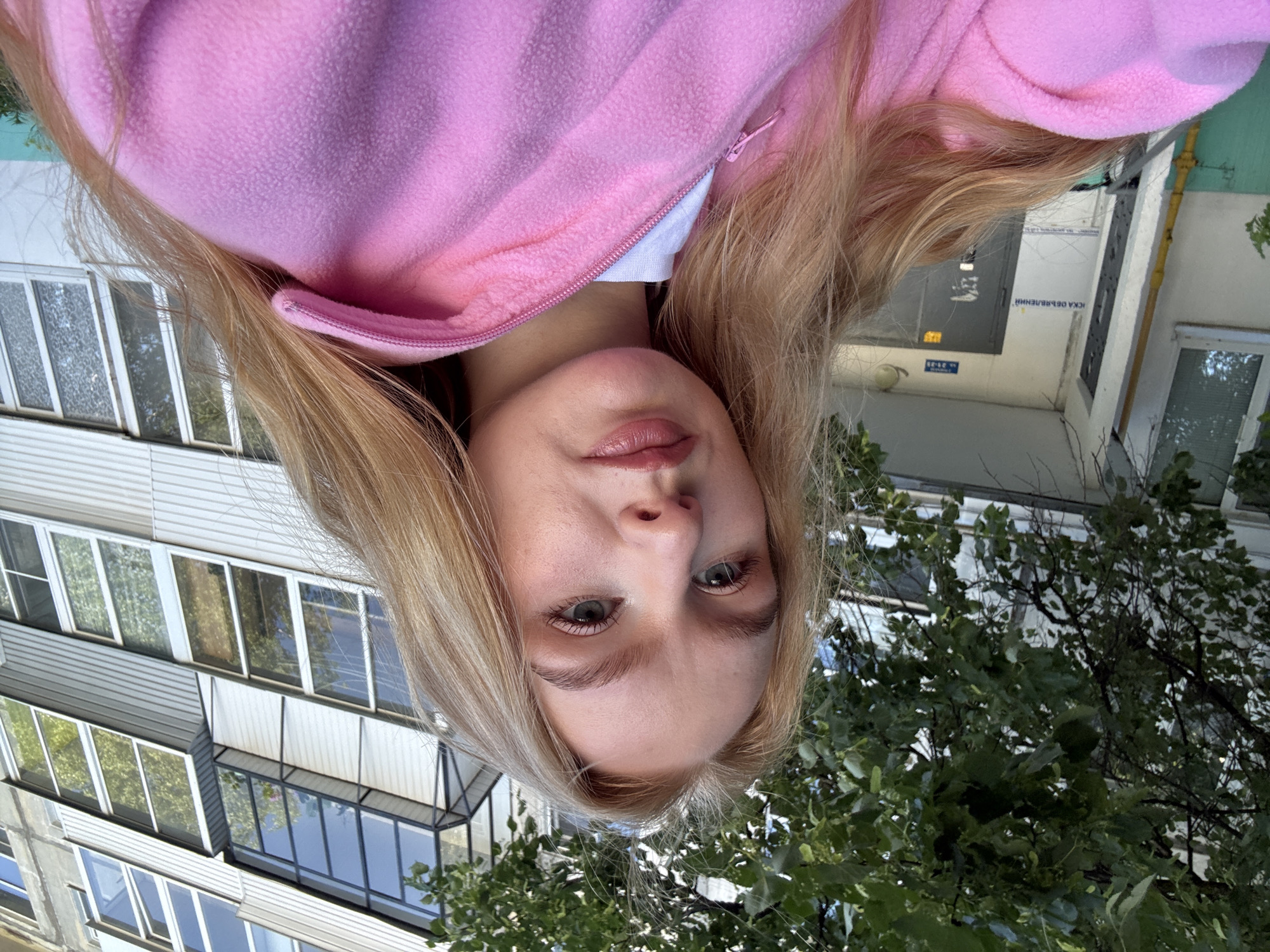 The image is a close-up selfie of a young woman with blonde hair, wearing a pink fleece jacket over a white t-shirt. She is standing outdoors in front of a multi-story apartment building. The building has several balconies with glass windows. Green trees are visible in the upper left corner of the image, partially obscuring the building. The woman is looking directly at the camera with a neutral expression. The scene appears to be set in the city of Chaikovsky, Russia, based on the building's architecture and a sign on the building that reads "ул. Западная 21-35" which translates to "Western Street 21-35."