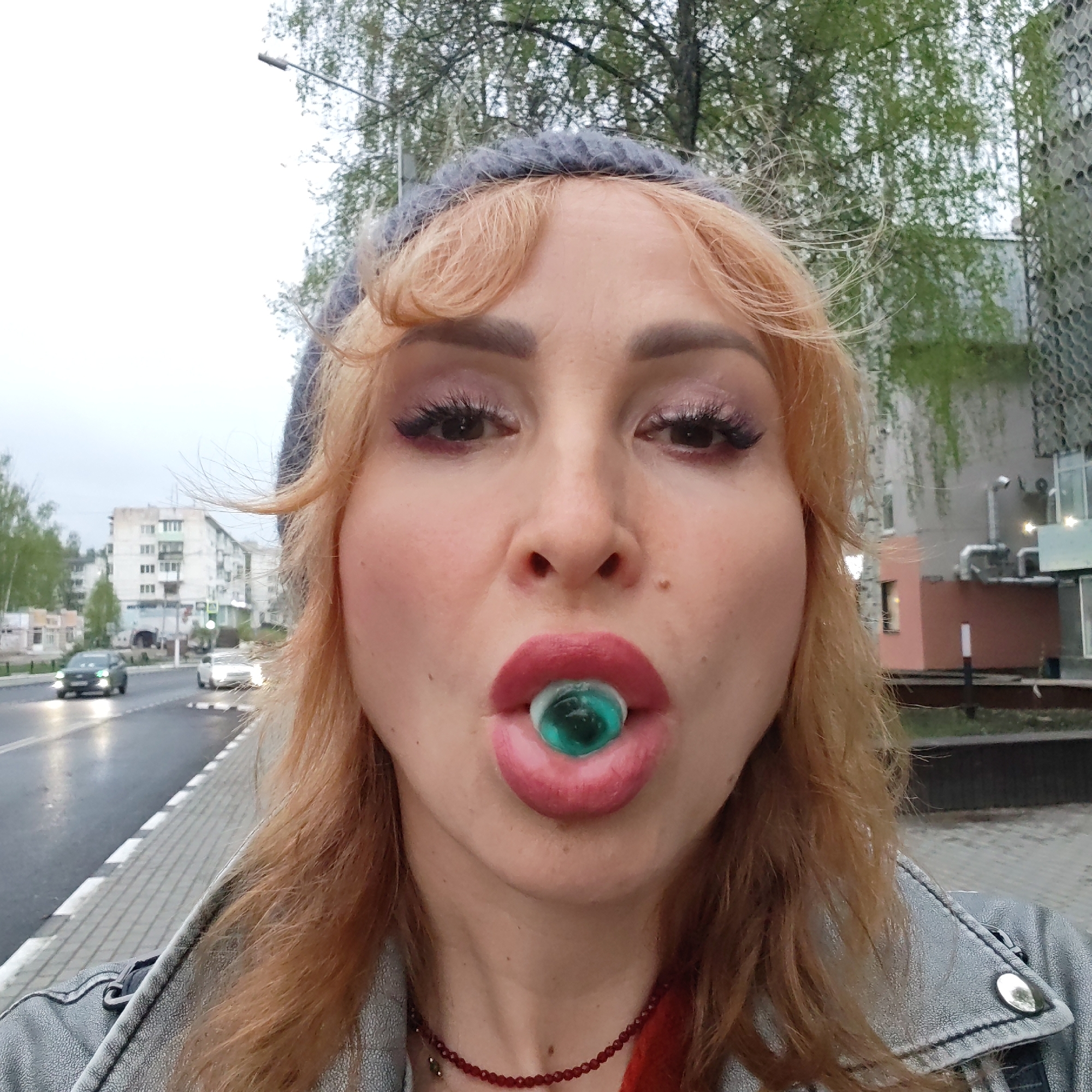 A woman with light red hair, wearing a gray knit hat and a denim jacket, is holding a green spherical object in her mouth. She is wearing makeup. She is on a sidewalk next to a street with cars, in Chaikovsky, Russia. Buildings and trees are visible in the background.