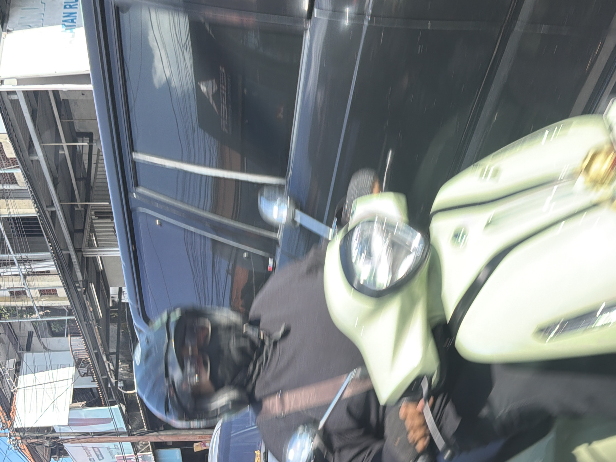 The image, rotated 90 degrees counter-clockwise, captures a street scene in bright daylight. In the foreground, a person wearing a black helmet and a black long-sleeved top is situated on a light green (possibly lime green) scooter. Their hands are on the handlebars, and a strap, likely from a bag, crosses their chest. The scooter's headlight and rearview mirrors are clearly visible, with some motion blur suggesting movement or recent stopping.
Adjacent to the scooter, a dark-colored SUV or large car occupies a significant portion of the frame. Its reflective surface shows a clear sky and environmental reflections. Faintly visible as a reflection on the car's window is the word "GIBRAN."
The background reveals a bustling urban or semi-urban environment with multiple buildings, overhead power lines, and what appears to be an elevated structure or scaffolding. A white banner with blue text is partially visible on one of the structures, displaying the fragment "ATAN RU". The bright, direct lighting indicates that the scene takes place during the day under clear weather conditions, in an active street setting consistent with North Kuta, Indonesia.