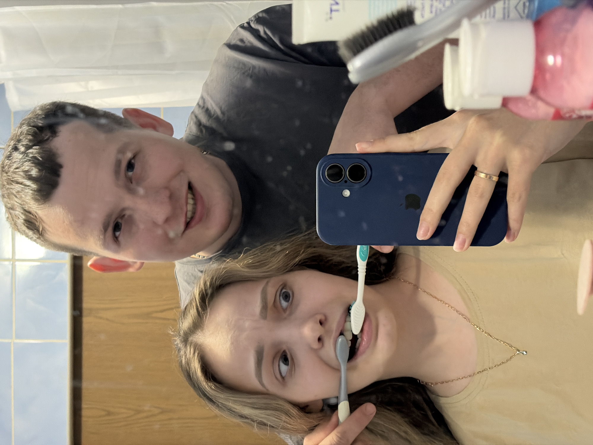 A selfie photo depicts a couple in a bathroom, presumably in the city of Chaikovsky, Russia. The man is partially visible in the upper left, smiling at the camera. The woman is holding a blue smartphone to take the picture. She is brushing her teeth with a grey toothbrush and a teal toothbrush in her mouth. She wears a gold necklace and a wedding band on her left ring finger. Various personal care items like a hairbrush, toothpaste tube, and a pink bottle are visible above the phone. The bathroom has wooden paneling and blue tiles on the walls.