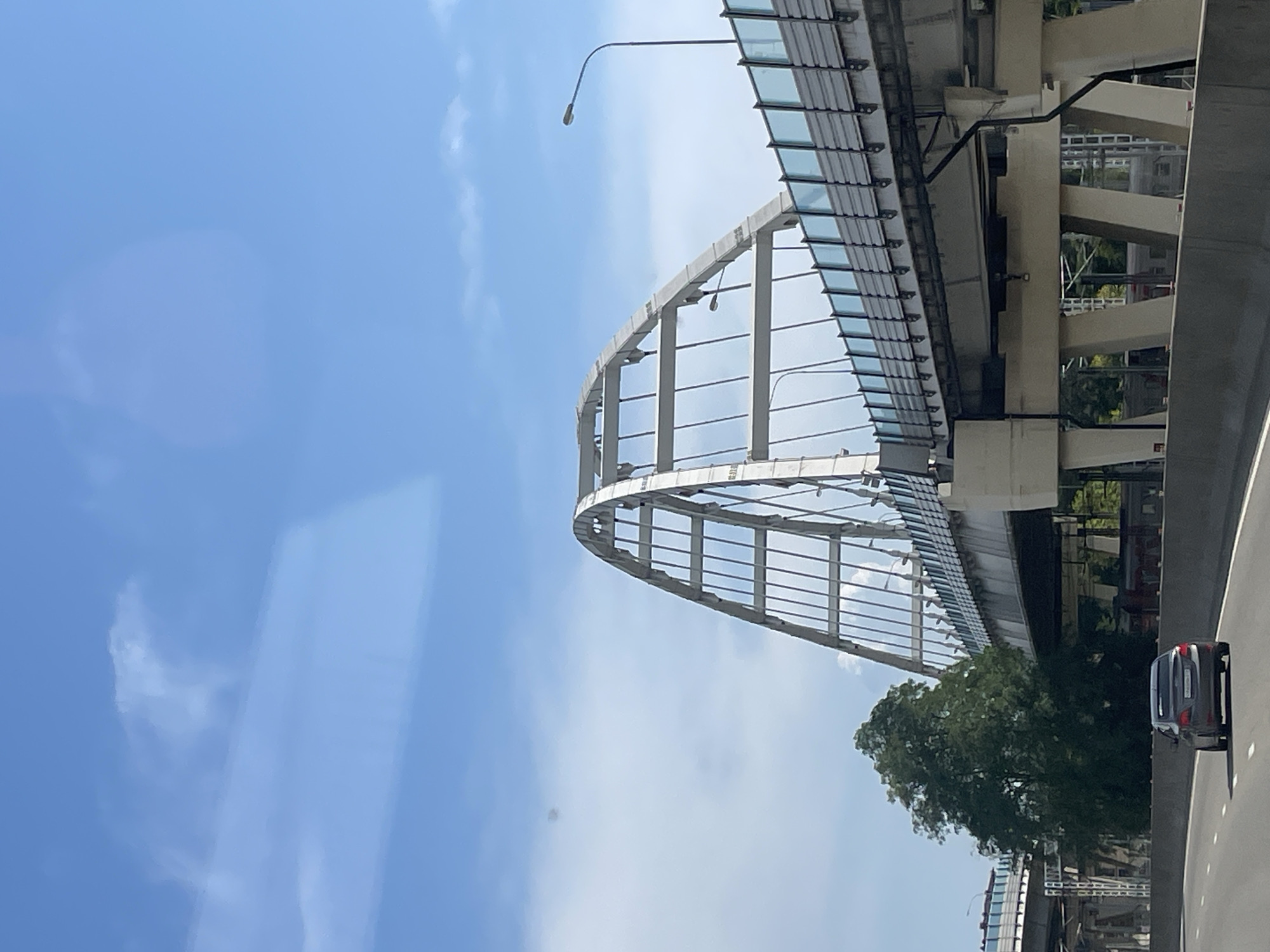 The image captures a view of a highway overpass and an arched bridge on a sunny day in Sochi, Russia. In the foreground, a dark-colored sedan is driving on the asphalt road. A large, dense green tree stands to the left of the road, partially obscuring a section of the bridge structure.
The main subject of the image is the large arched bridge, which is white and made of a metal lattice structure. The bridge spans across the highway, with multiple lanes of traffic visible on its upper level. The railings on the upper bridge have a blue translucent material. A street lamp is visible on the right side of the upper bridge.
In the background, under the bridge, there are glimpses of what appear to be train tracks and a train. The sky above is a bright blue with scattered white clouds. The overall impression is of a modern infrastructure scene within a city, likely a transportation hub given the presence of both a highway and a train. The specific location is the Arched Bridge over the Mzymta River in Sochi, Russia, known for its distinctive design and importance as a transportation artery.