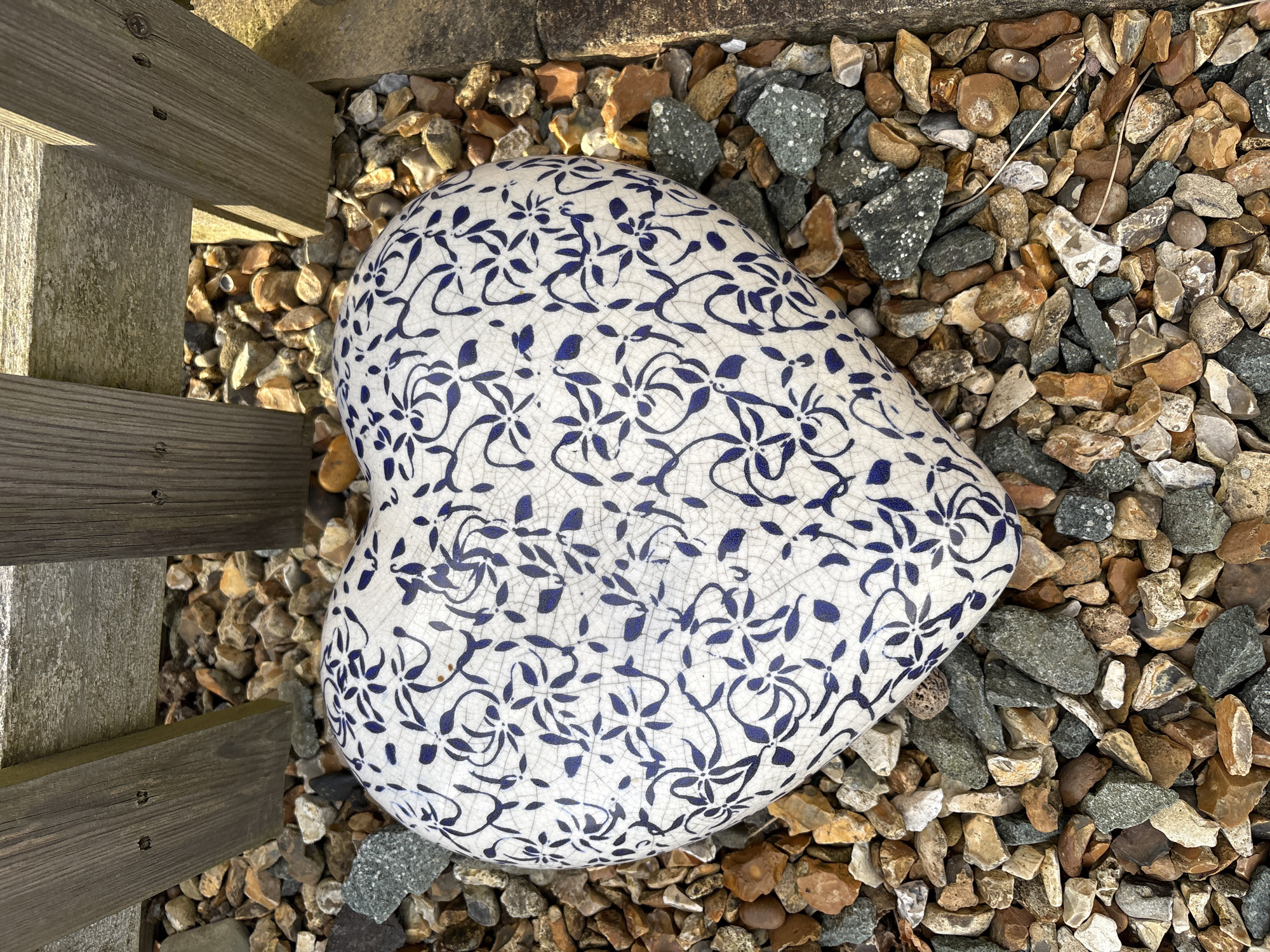 The image depicts an outdoor scene at ground level in Stiperstones, United Kingdom. A heart-shaped ceramic object is centrally positioned, resting on a bed of multicolored gravel. The object is white with a repeating blue floral pattern and features a crackle glaze finish. The gravel consists of irregularly shaped stones in shades of brown, grey, and white. In the upper background, two weathered wooden planks, consistent with fence posts or a garden border, are partially visible, casting shadows onto the gravel. No individuals, animals, or active interactions are present within the frame.