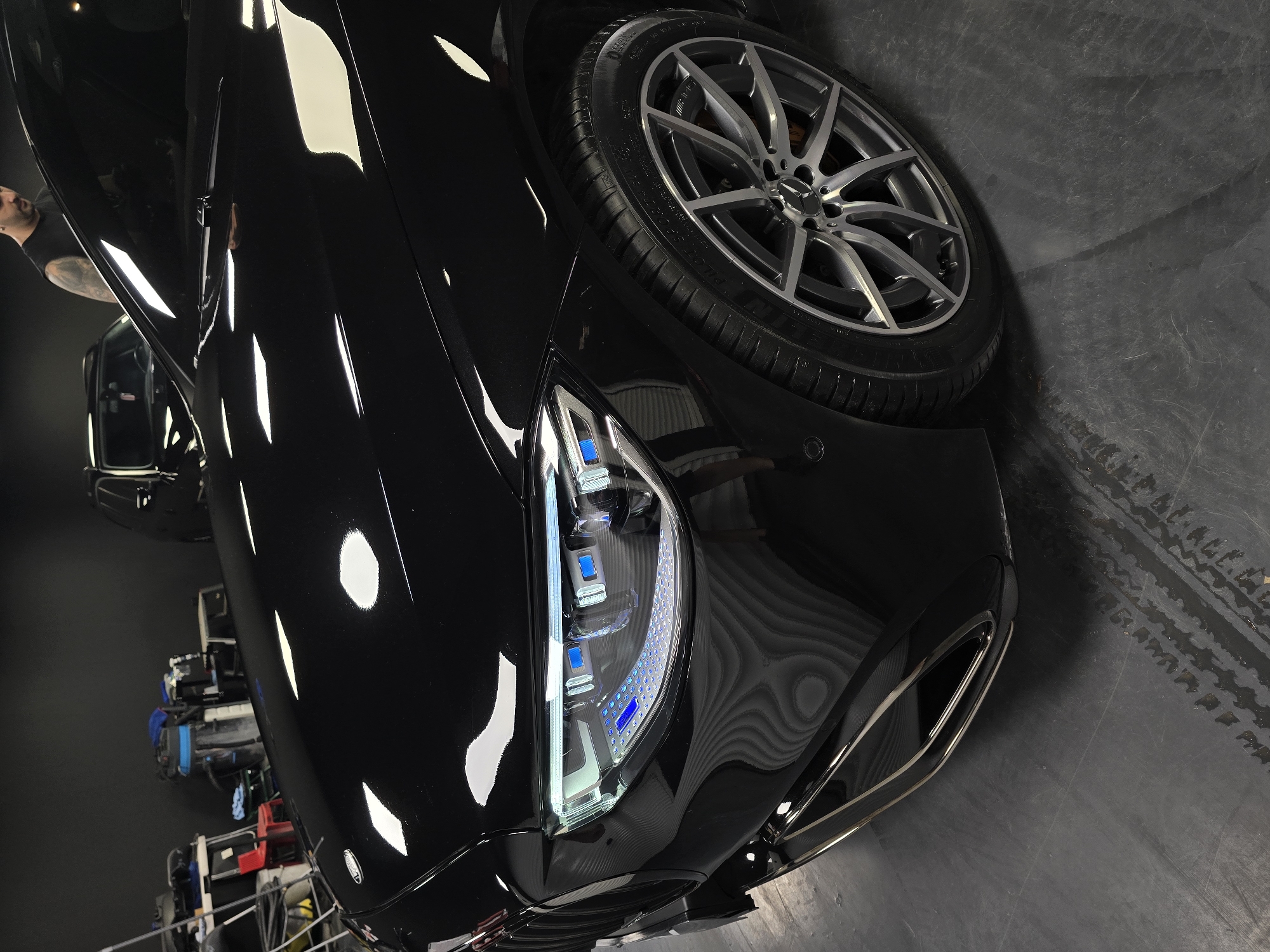 The image shows a close-up, low-angle view of the front left side of a black luxury car, likely a Mercedes-Benz S-Class, parked indoors. The car's glossy finish reflects the surrounding environment, including overhead lights and other objects in the background. The headlights of the car are illuminated, displaying a complex LED design with blue accents. The front wheel, with a multi-spoke alloy rim, is visible. In the background, shelves stocked with car cleaning supplies and a vacuum cleaner are present, suggesting the car is in a detailing or workshop setting. A dark-colored SUV is also partially visible in the far background, and a man with a tattooed arm and wearing a black sleeveless shirt is standing behind the car, out of focus. The floor appears to be a dark, possibly concrete surface with some wet tire marks. The location is not specified as Ruse, Bulgaria.