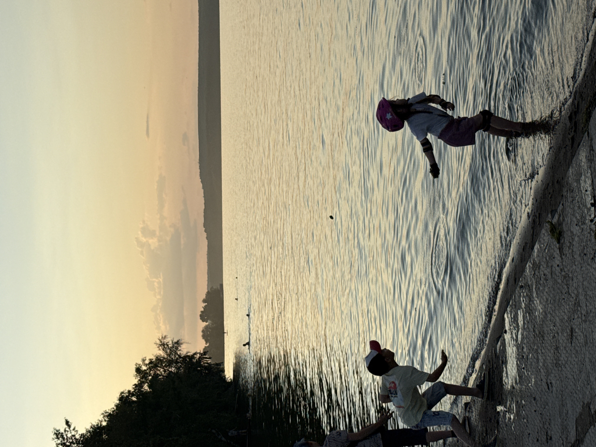 The image depicts two children near a body of water, likely Votkinsk, Russia, during a sunset. The scene is a shoreline where the water meets a paved or concrete area.
In the foreground, a young boy wearing a t-shirt, shorts, and a cap is looking upwards, his right arm extended as if reaching for something. He appears engaged in an activity or conversation with someone out of frame.
Further back, on the water's edge, a child in a helmet, t-shirt, and shorts is in motion. They seem to be rollerblading or skating, with their body leaning to one side, suggesting movement. Protective gear, including knee and elbow pads, is visible.
The water exhibits rippling patterns, reflecting the soft, warm light of the setting sun, creating a serene atmosphere. The sky transitions from a pale yellow near the horizon to a lighter hue higher up, with faint clouds present. Silhouetted trees and a distant landmass form the backdrop on the left side of the image. The overall mood is one of playful activity and natural beauty during the golden hour.