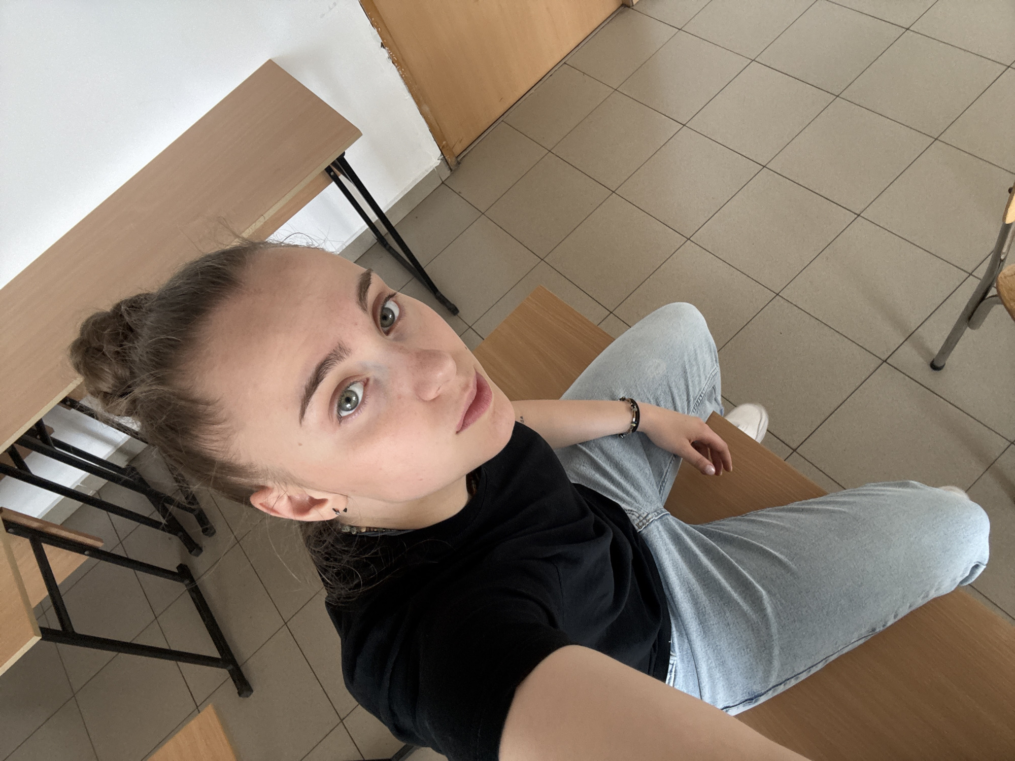 A young woman with light skin and green eyes sits on a wooden desk in what appears to be a classroom. She has her hair pulled back in a bun and wears a black t-shirt and light blue jeans. Her left hand is resting on her leg, and she is wearing a bracelet on her left wrist. The classroom has beige tile flooring and light-colored wooden desks with black metal legs. A white wall is visible on the left side of the image. The image has a slightly upward perspective. The location is uncertain without more information, and therefore I can't ascertain that it's in Sofia, Bulgaria.