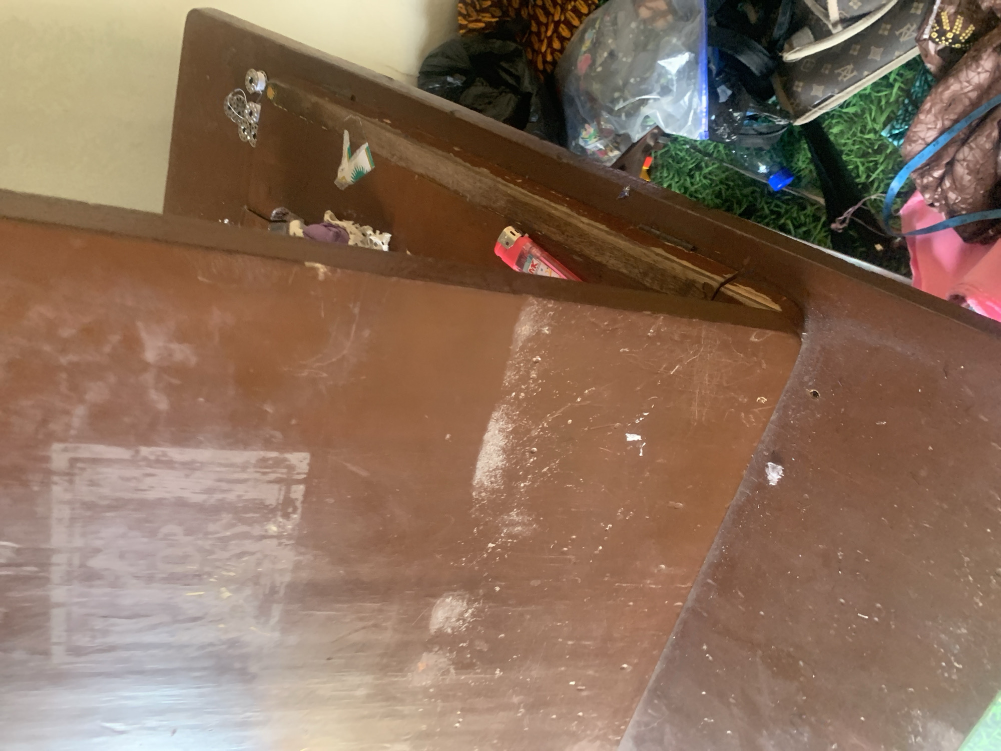 The image shows a close-up of a wooden desk with various objects on its surface and the floor below. The desk is a dark, reddish-brown color and appears to be dusty and scratched. On the desk surface, there are several small items, including what looks like a pink lighter, a spool of thread, and some decorative metal findings. In the background, a black trash bag is visible, along with other assorted items. The floor beneath the desk is covered with green material that resembles artificial turf, and more bags and fabric are scattered around. The overall impression is one of a cluttered and somewhat neglected workspace.
The location of Jauro Yinu, Nigeria, does not appear to be relevant to the visual content of the image, as there are no specific landmarks or contextual clues that indicate this particular geographic setting.