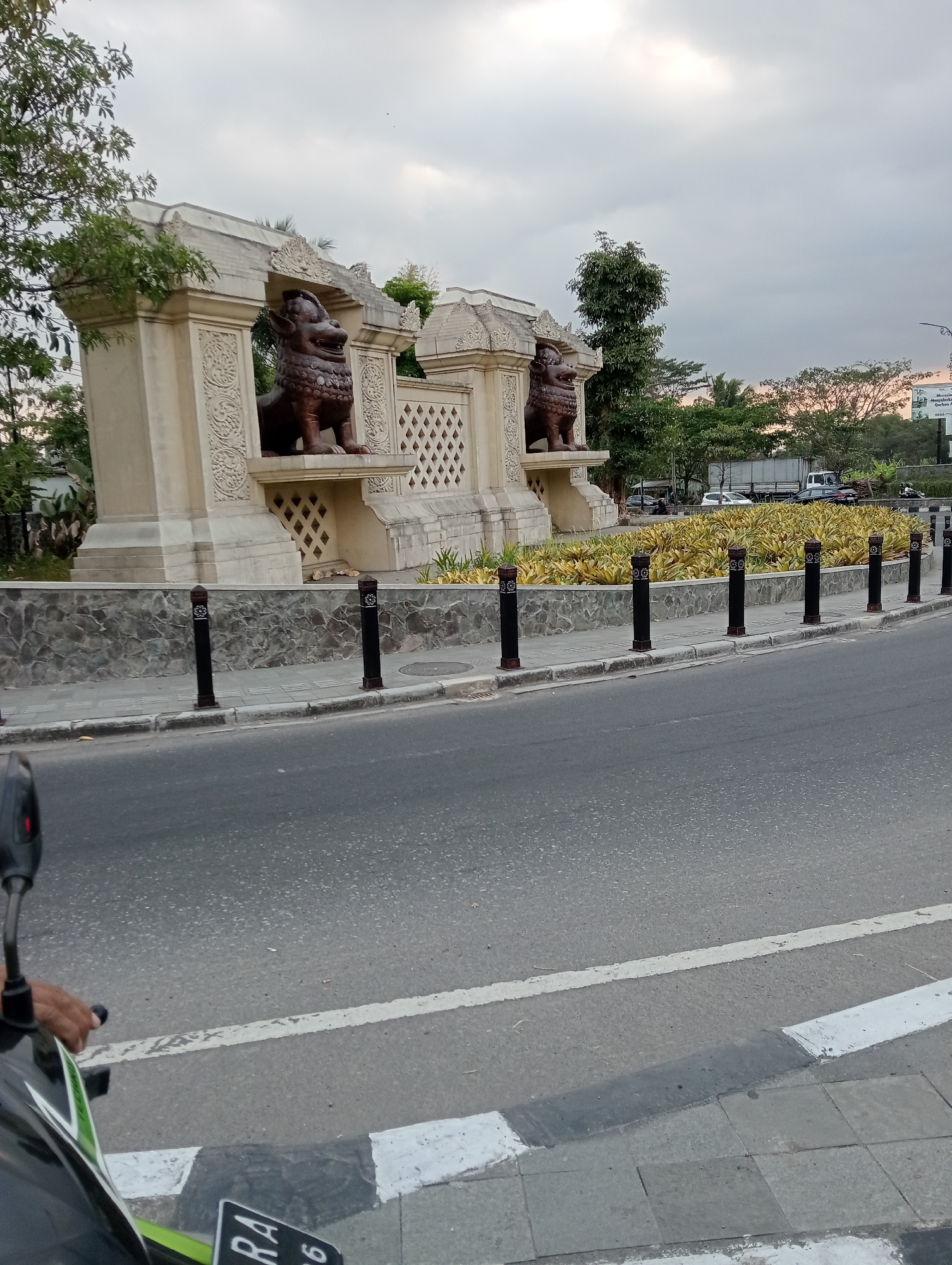 In the city of Pabelan, Indonesia, a street view captures a prominent monument with two stylized lion statues positioned within archways. The monument is constructed of light-colored stone with intricate carvings on its facade. A low stone wall separates the monument from a sidewalk adorned with black bollards topped with decorative emblems. Beyond the sidewalk, a paved road curves to the right, with white and black markings indicating a pedestrian crossing. The scene is framed by trees and vegetation, and the sky overhead is overcast. A portion of a motorcycle, including a hand and rearview mirror, is visible in the foreground, suggesting the image was taken from a moving vehicle. In the background, vehicles, including trucks and cars, are visible, along with some buildings and distant trees.