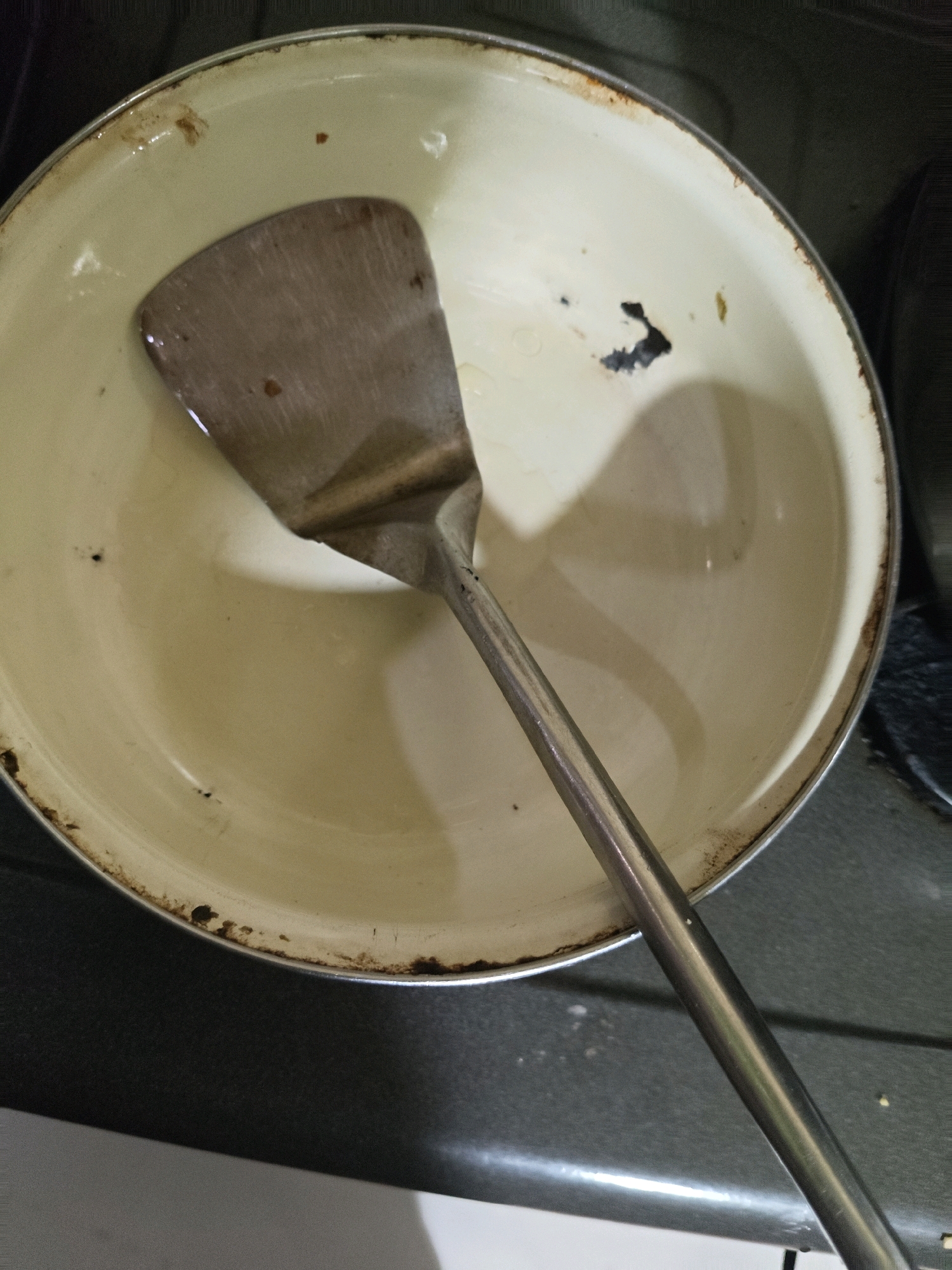 A metal spatula rests inside a shallow, light-colored pot with visible rust and stains along the rim. The pot appears to be on a dark-colored surface, possibly a stovetop. The lighting is dim, casting shadows within the pot. The image was taken in Sumberwono, Indonesia.