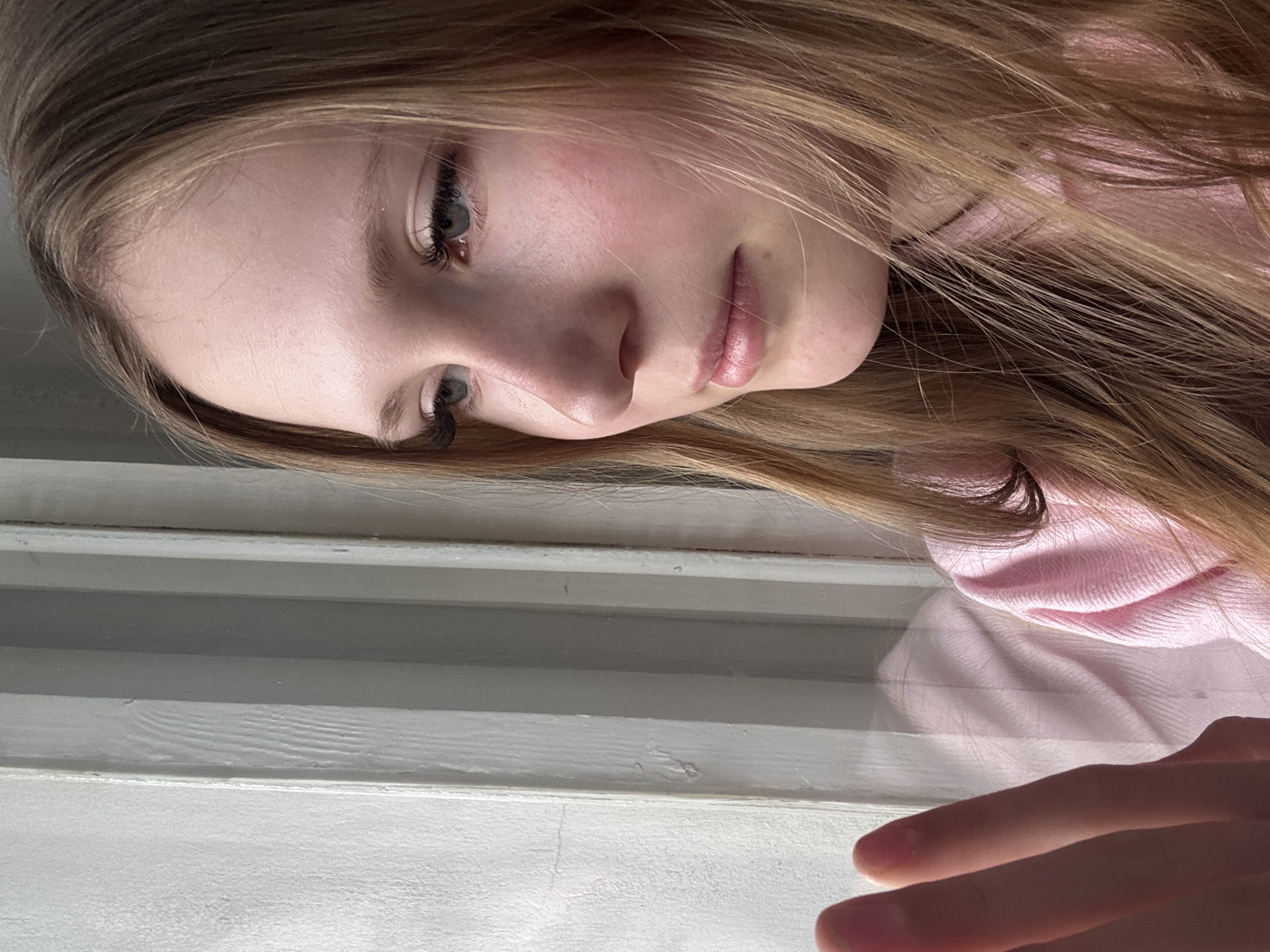 A young person with light skin and light brown hair is looking down at the camera. The subject is wearing a pink top and has fair features. The person is positioned above what appears to be a white-painted window frame or another light-colored horizontal structure. A hand with visible fingers is positioned in the lower right corner. The location in the city of Chaikovsky, Russia, cannot be confirmed from the visual information provided.