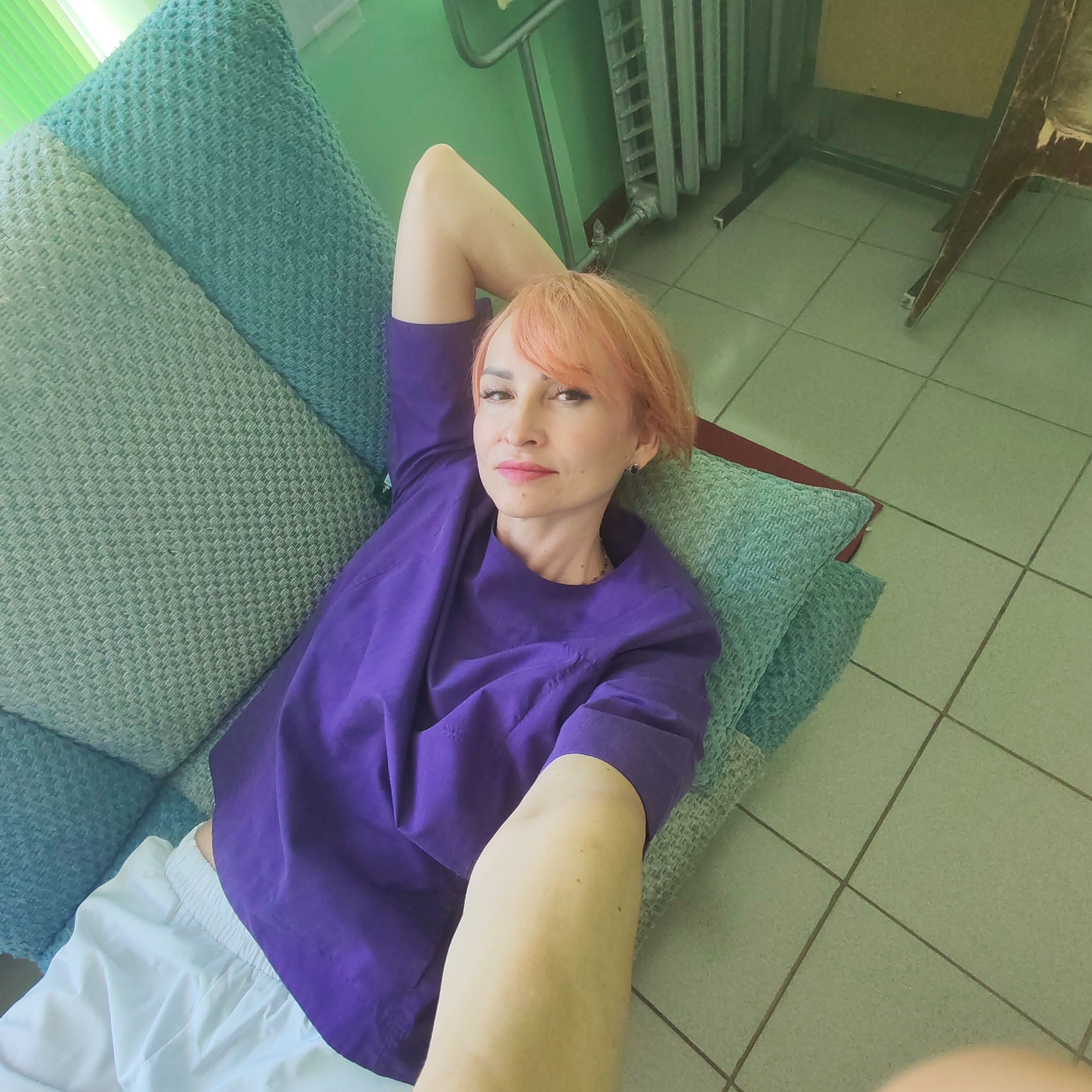 A woman with short, light pink hair is depicted lying on her back on a sofa or daybed, looking directly at the camera with a soft smile. She is wearing a purple short-sleeved top and white pants or shorts. Her left arm is extended towards the camera, indicating she is taking a selfie.
She is supported by two textured cushions, one in teal/light blue and the other in a lighter green shade. The background features a light green wall. On the right side of the frame, a dark grey radiator or heating unit is visible against the wall, along with the lower portion of a wooden piece of furniture, possibly a chair or desk. The floor is covered with light green square tiles with dark grout lines.
The scene takes place indoors, likely during the daytime, given the natural light present. The woman appears relaxed and content. The setting has a somewhat utilitarian or older aesthetic, suggested by the radiator and tiled floor. No text is visible in the image. The location is Chaikovsky, Russia.