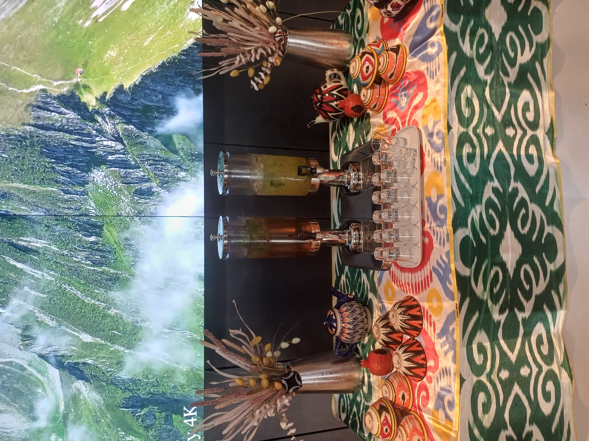 The image displays a table set for a drink service. In the background, a large screen shows a scenic view of green mountains with mist and a small red building on a ridge. In the foreground, two large glass beverage dispensers are positioned side-by-side on a black stand. The left dispenser contains a dark amber liquid, likely tea, and the right dispenser holds a lighter, greenish liquid with ice and mint. To the left of the dispensers, a tall, metallic vase holds a dried floral arrangement. In front of the vase, a small, ornate blue and white teapot sits on a colorful, patterned tablecloth. Next to the teapot, there is a single red pomegranate. A collection of small, decorative bowls with intricate patterns in red, yellow, and black are arranged on the tablecloth. To the right of the dispensers, a tray holds several clear shot glasses. The overall scene suggests a refreshment station, possibly in a hotel or event venue. There are no people visible in the image. The location is identified as the city of Myk, Uzbekistan, implied by the text overlay on the screen, though the image itself does not provide explicit geographical markers beyond that.
