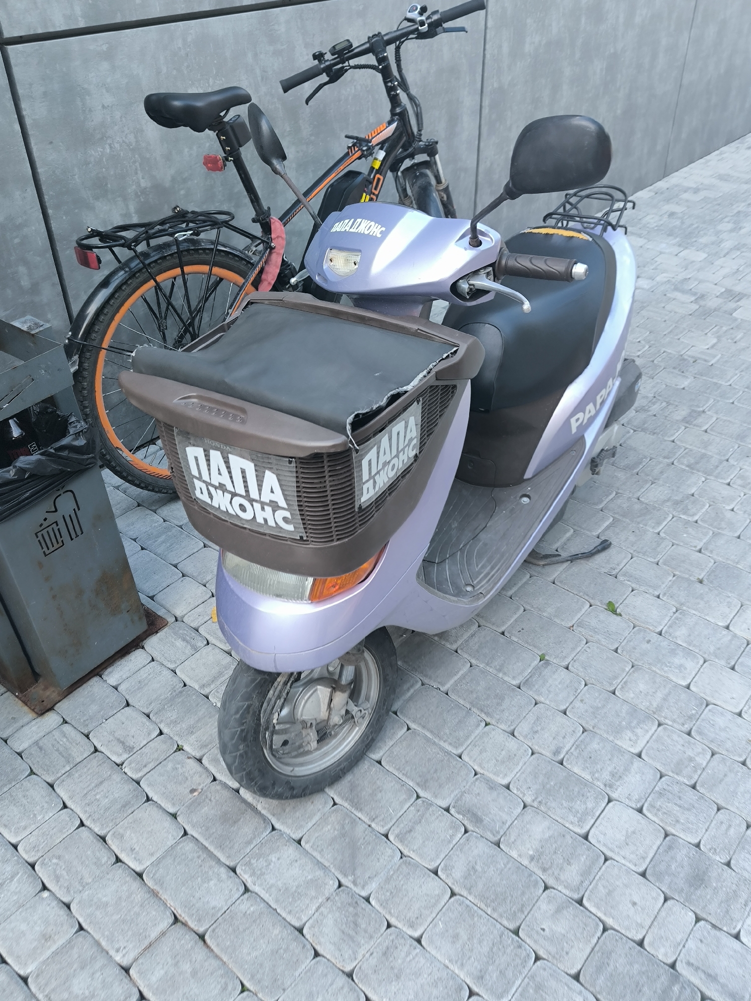 The image shows a purple scooter and a bicycle parked on a sidewalk made of rectangular pavers. A grey metal trash bin is to the left of the scooter. Both vehicles have white lettering on them. The scooter has "PAPA ДЖОНС" on its front basket and "PAPA" on its side. The bicycle has "Papa" visible on its frame. The background is a textured grey wall. The scene is outdoors, likely in a city, but there are no explicit indicators of Novosibirsk, Russia.