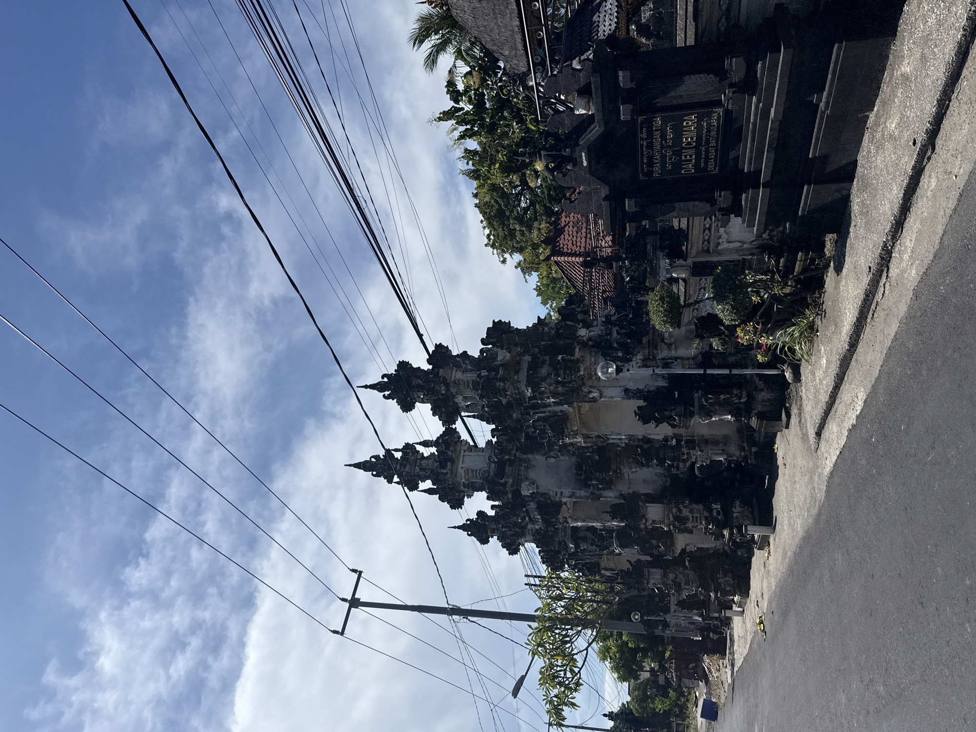 The image depicts a street scene in Batununggal, Indonesia. On the right is a section of a traditional Balinese wall adorned with intricate carvings and a sign in Indonesian, "Pura Kahyangan Tiga Dalem Cemara Desa Adat Batumulapan." The wall borders the street. To the left, power lines run diagonally across the sky, which is partly cloudy. A utility pole stands near the road. The street appears to be paved. There is no indication of people in the scene. The focus is on the architectural detail and the Balinese aesthetic.