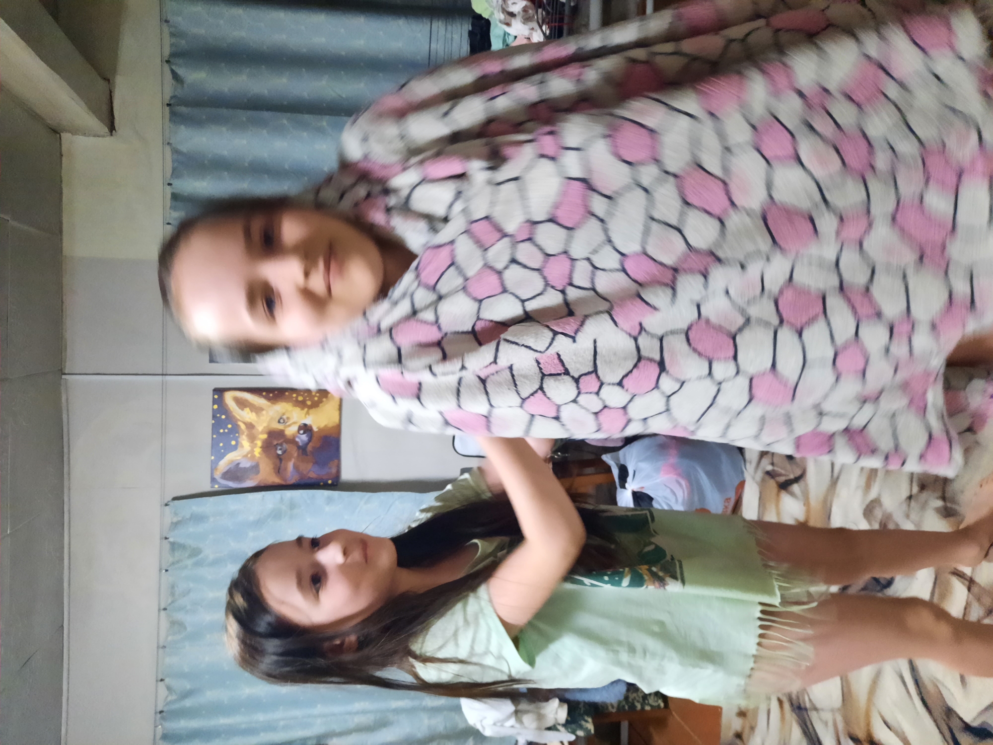 Two girls are depicted indoors. One girl is standing and wearing a light green t-shirt. The other girl is wrapped in a patterned blanket with pink and white designs. The setting appears to be a bedroom, with a painting of a fox visible on the wall and patterned curtains. The location is Krasnogorskoye, Russia.