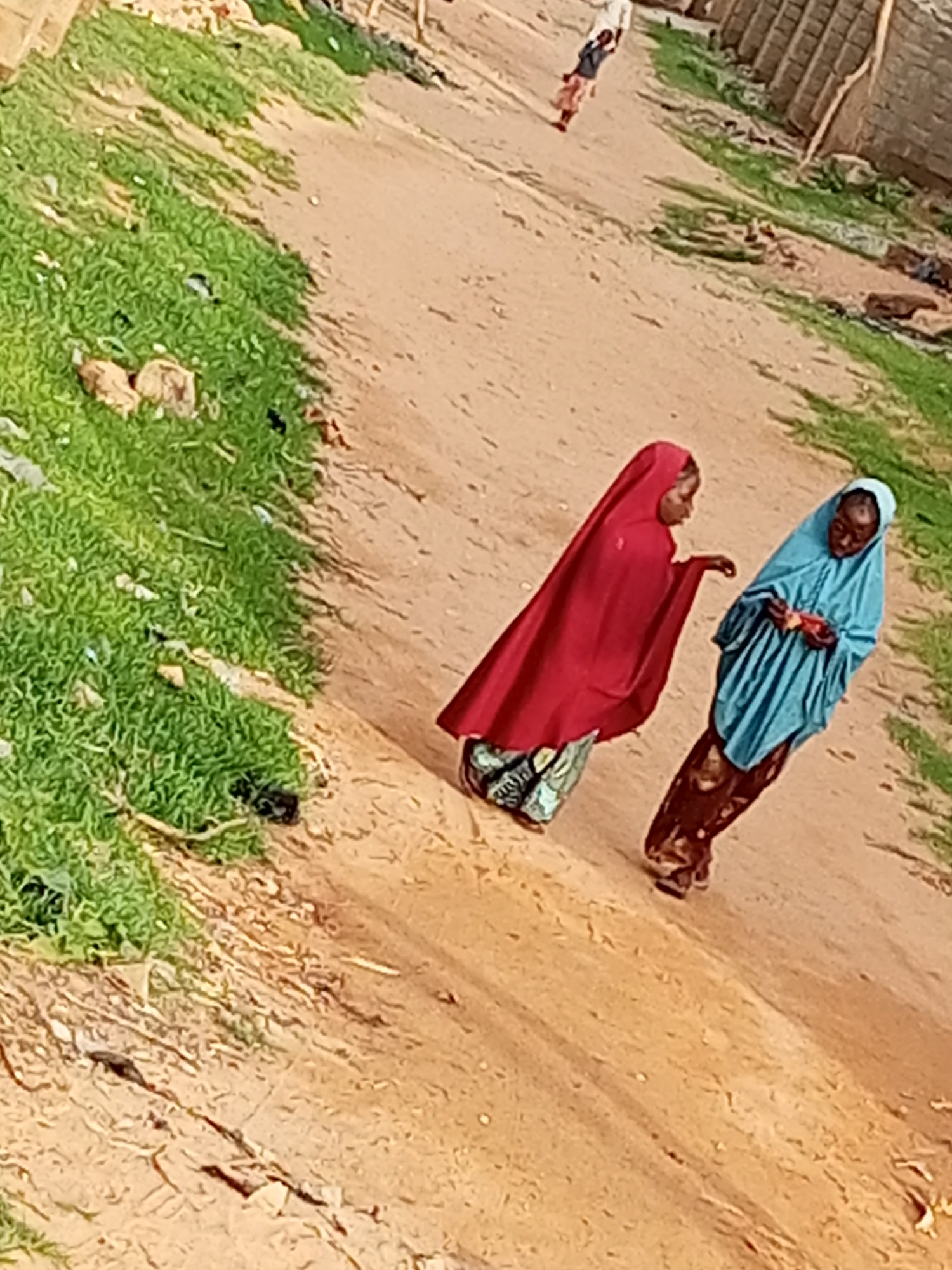 Two young women are standing on a dirt path in Tempure, Nigeria. The woman on the left is wearing a red headscarf and a patterned skirt. The woman on the right is wearing a blue headscarf and a patterned skirt. The path is flanked by dry grass and some debris. In the background, there are some simple brick structures and a further scattering of grass. The sky is not visible. The image is taken from a slightly elevated, oblique angle.