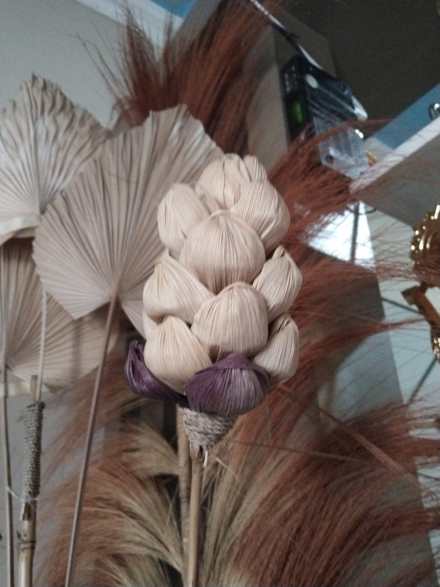 The image displays a close-up of dried decorative plant materials, likely arranged as an indoor floral display. A cluster of light-colored, bulbous elements, resembling dried flower heads or seed pods, is prominently featured in the center. These elements are gathered together and bound by a band of rough, woven material, possibly twine or straw. Beneath this cluster, a smaller section of darker, purplish material is visible, also appearing to be part of the dried plant. Surrounding this central arrangement are feathery, dried fronds and grasses in various shades of brown and tan, creating a naturalistic and textured backdrop. To the left, a large, fan-shaped dried leaf or palm frond is partially visible, its ribbed structure contributing to the overall organic aesthetic. In the background, slightly out of focus, is a portion of an electronic device, possibly a router or modem, mounted on a wall. The overall impression is one of rustic, dried botanical decor. The image does not contain any discernable elements that would allow for the identification of its specific location within Tasikmalaya, Indonesia.