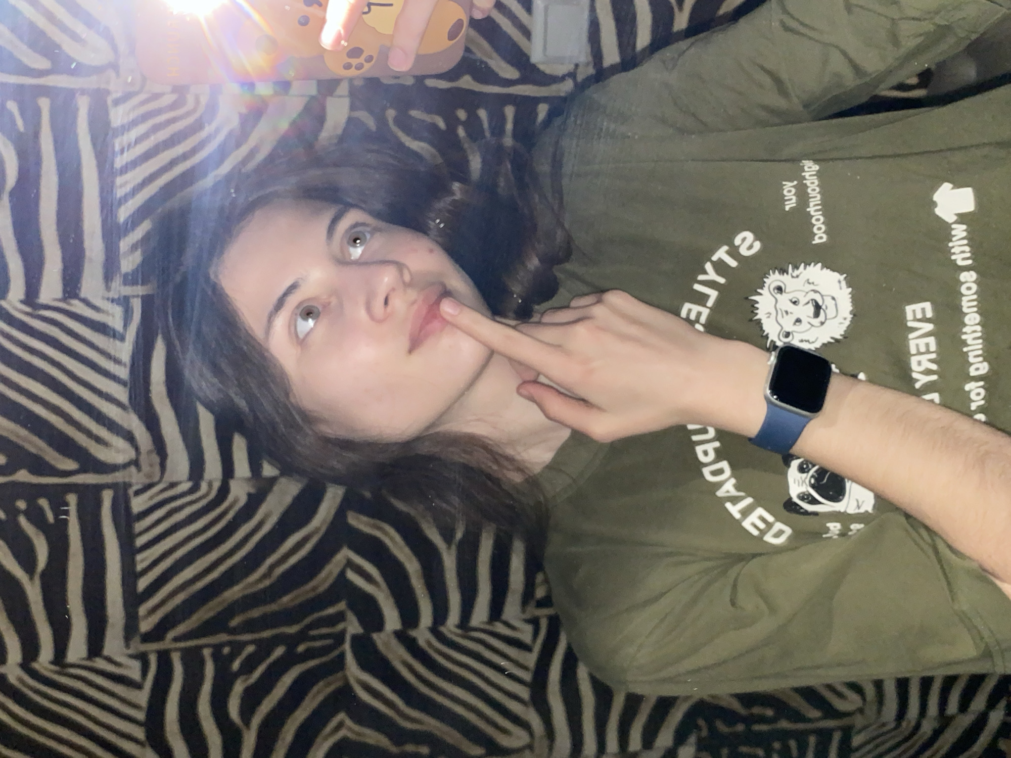 A young person with light skin lies on a zebra-striped surface, likely a bed or couch. They wear a green long-sleeved shirt with various cartoonish animal designs and text. A blue smartwatch is visible on their left wrist. The person's left hand is raised, with the index finger touching their lips. The person has dark hair and is looking upwards. A bright light source obscures the top portion of the image.
There is no information to suggest the location is Votkinsk, Russia.