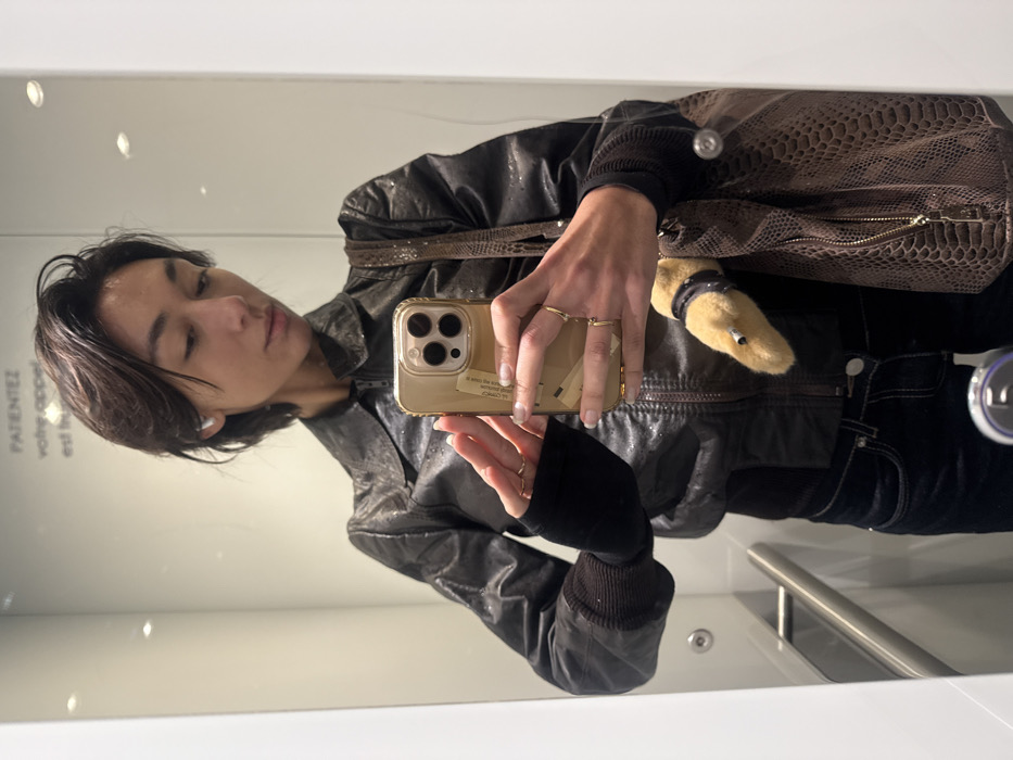 A person with dark, short hair, wearing a black bomber jacket with ribbed cuffs, a dark inner garment, and dark jeans, is depicted in a mirror selfie. A white earbud is visible in their left ear. They hold a gold-colored smartphone with a prominent camera module and a label affixed to its back in their right hand, displaying multiple thin gold-toned rings on their fingers. A brown, textured bag, appearing to be snakeskin-patterned, hangs on their left side, with a light brown plush animal head (possibly a duck) with a dark collar peeking out from behind it. The reflection reveals a brightly lit, modern indoor environment with white walls and a metallic horizontal handrail. French text, partially visible as "PATIENTEZ votre appel est...", is printed on the mirrored surface, indicating a waiting area or reception within a public or private facility in Paris, France.