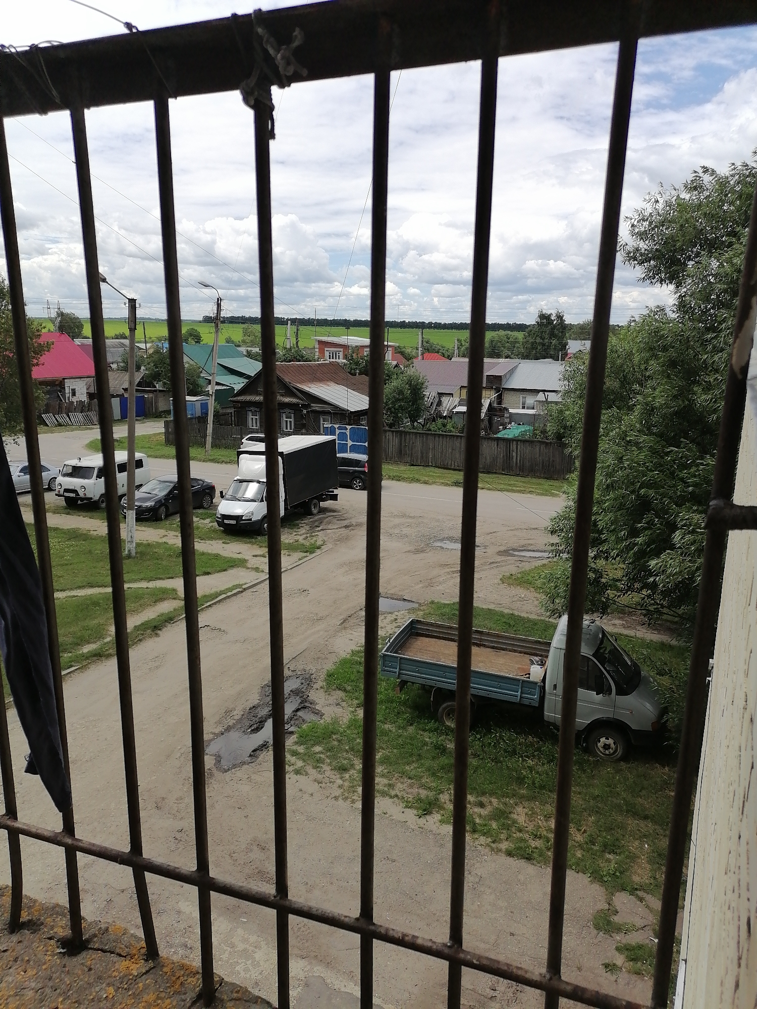 This is a high-angle, full shot taken from behind metal bars, looking out at a residential street scene in the city of Kuznetsk, Russia. The foreground is dominated by the rusty, dark metal bars of a window or balcony railing. Beyond the bars, a dirt and gravel road curves through a village-like setting. On the left side of the road, parked are a white van and a dark sedan. Further down the road on the left, a white box truck is parked. On the right side of the road, a light-colored flatbed truck is parked on a grassy area, partially obscured by trees. The background features several houses with various roof colors, including red, green, and brown. Beyond the houses, there are fields of green crops and a cloudy sky. The overall impression is of a rural or semi-rural residential area.