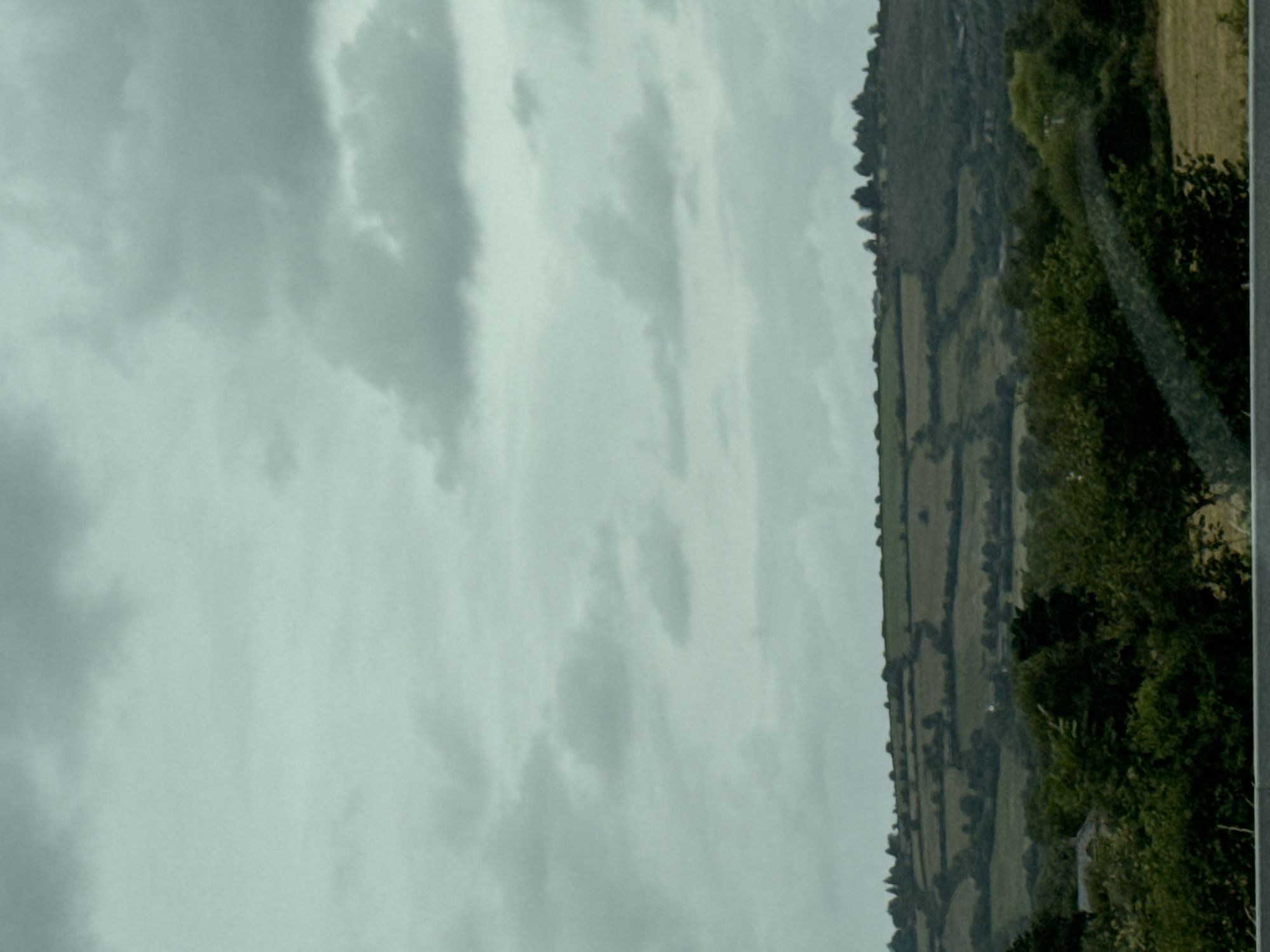 The image depicts a rural landscape under a cloudy sky. Rolling hills covered in green fields and hedgerows dominate the middle ground, suggesting agricultural land. A dark line of trees runs along the crest of the hills. In the foreground, a dense area of dark green trees partially obscures a lighter green field. The sky is filled with numerous gray clouds, indicating an overcast day. No people or specific actions are visible. The location is identified as Craan, Ireland.