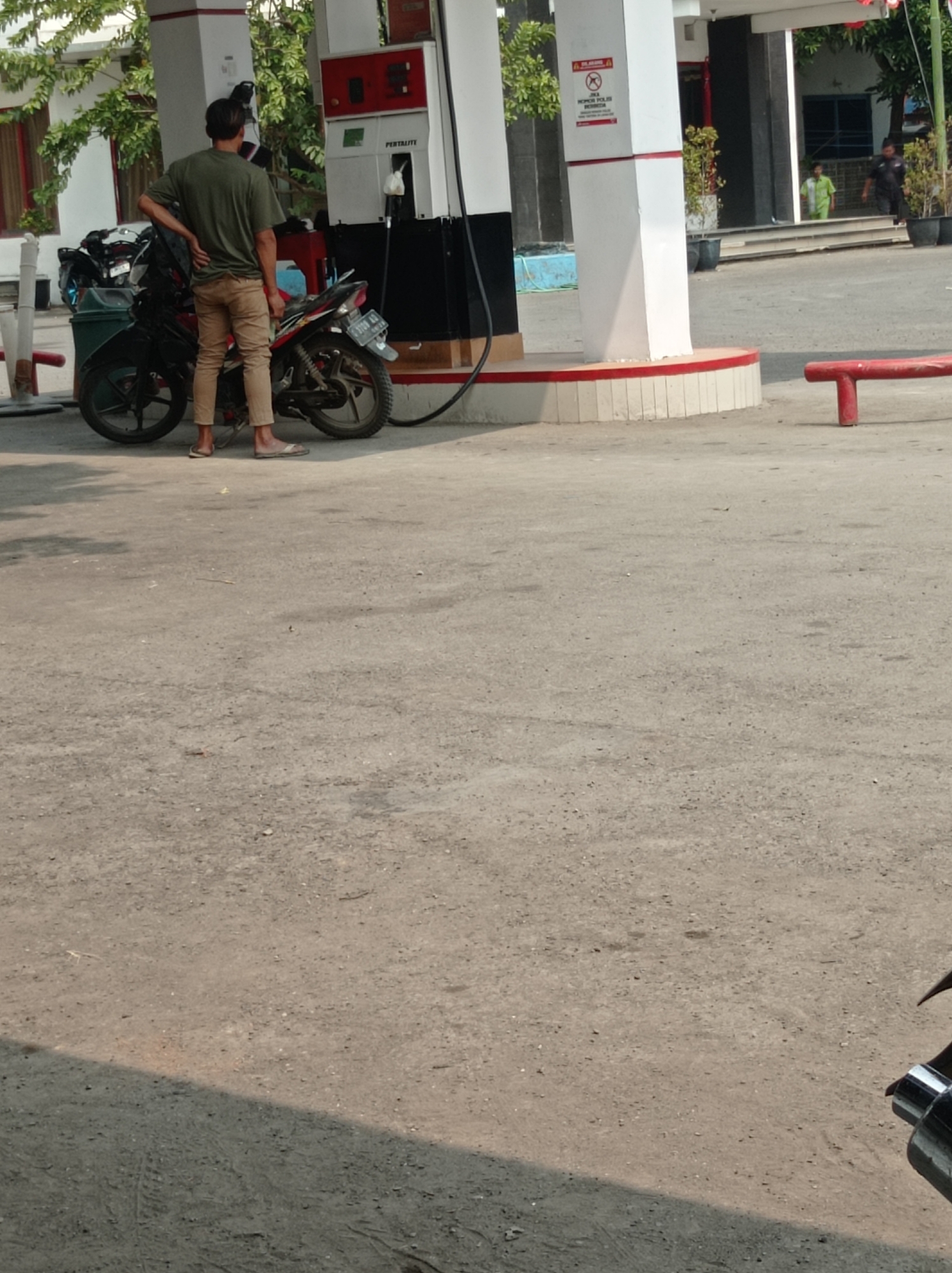 A lone individual, wearing a green t-shirt and beige pants, stands next to a red and black motorcycle at a Pertamina gas station in Kramat, Indonesia. The motorcycle is parked in front of a gas pump, and the individual is looking away from the camera, with their left hand resting on their hip. Behind the individual, a building with white and red pillars and a dark-colored entrance is visible. Two other individuals can be seen in the background, one wearing a light green uniform and another in dark clothing. The ground is paved with asphalt, and the scene is illuminated by sunlight, casting shadows on the pavement.