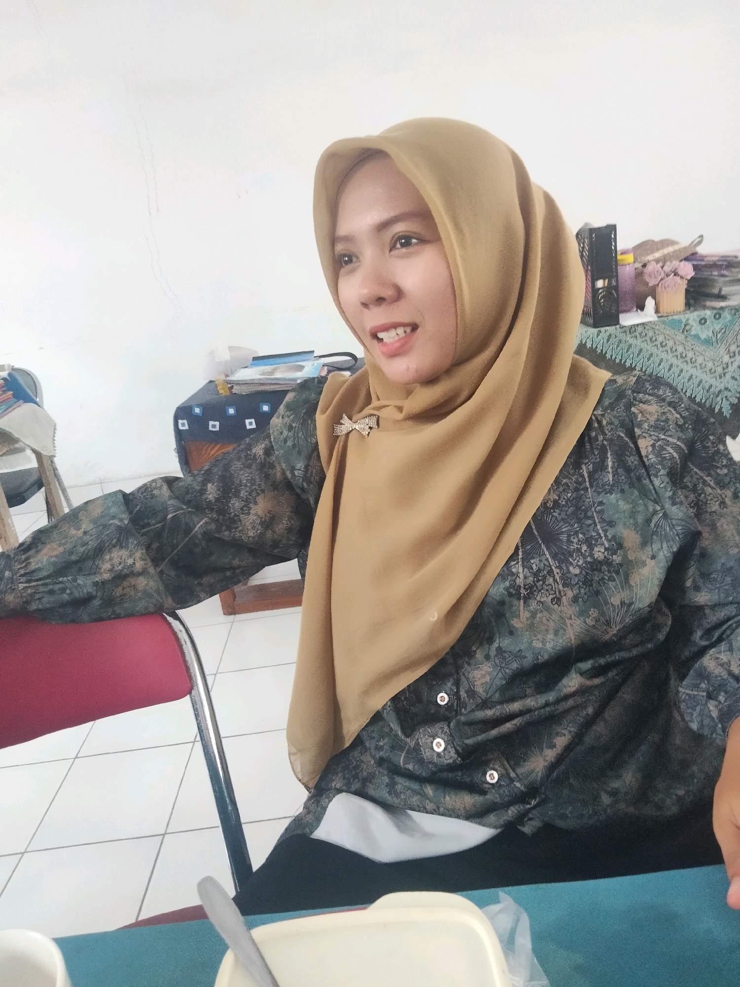A woman wearing a tan hijab and a dark, patterned shirt with a brooch is seated at a table in what appears to be a room with a white tiled floor. The room has a white wall and a table covered with a patterned cloth in the background, on which are several items including a black basket, some bottles, and a small vase with pink flowers. The woman is looking to her left with a slight smile. A red chair is visible to the left of the woman, and a blue tablecloth is on the table in front of her, with a white cup and a spoon on it. The overall impression is of a casual indoor setting, likely within the city of Suka Sari, Indonesia, based on the contextual information provided.