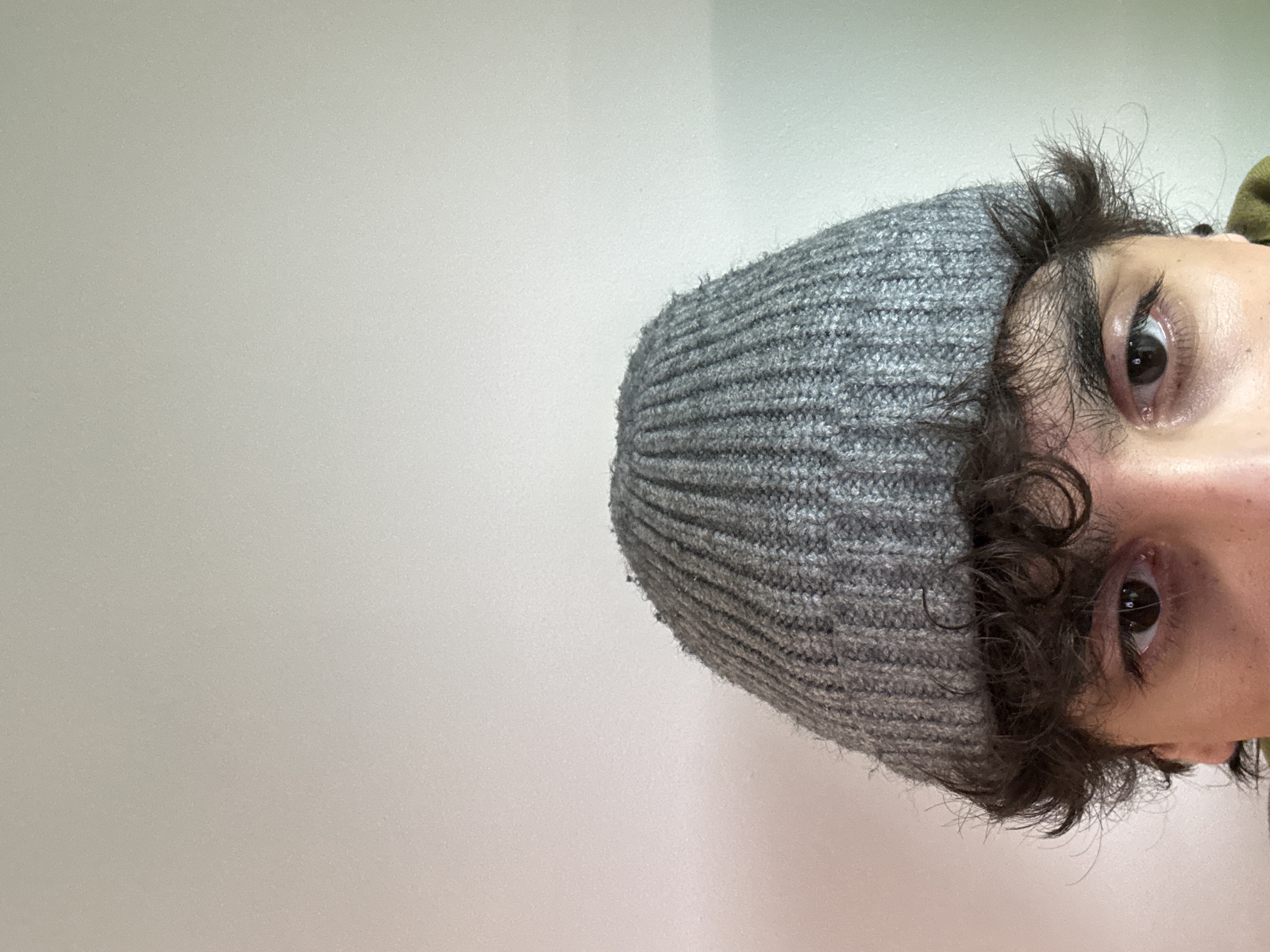 The image shows a close-up of a person's face, rotated sideways. The person is wearing a grey knitted beanie. Visible features include dark curly hair peeking out from under the beanie, two eyes with dark irises, and portions of the face and ear. The background is a plain, light-colored wall. There is no indication of Moscow, Russia, in the image. No actions or interactions are depicted.