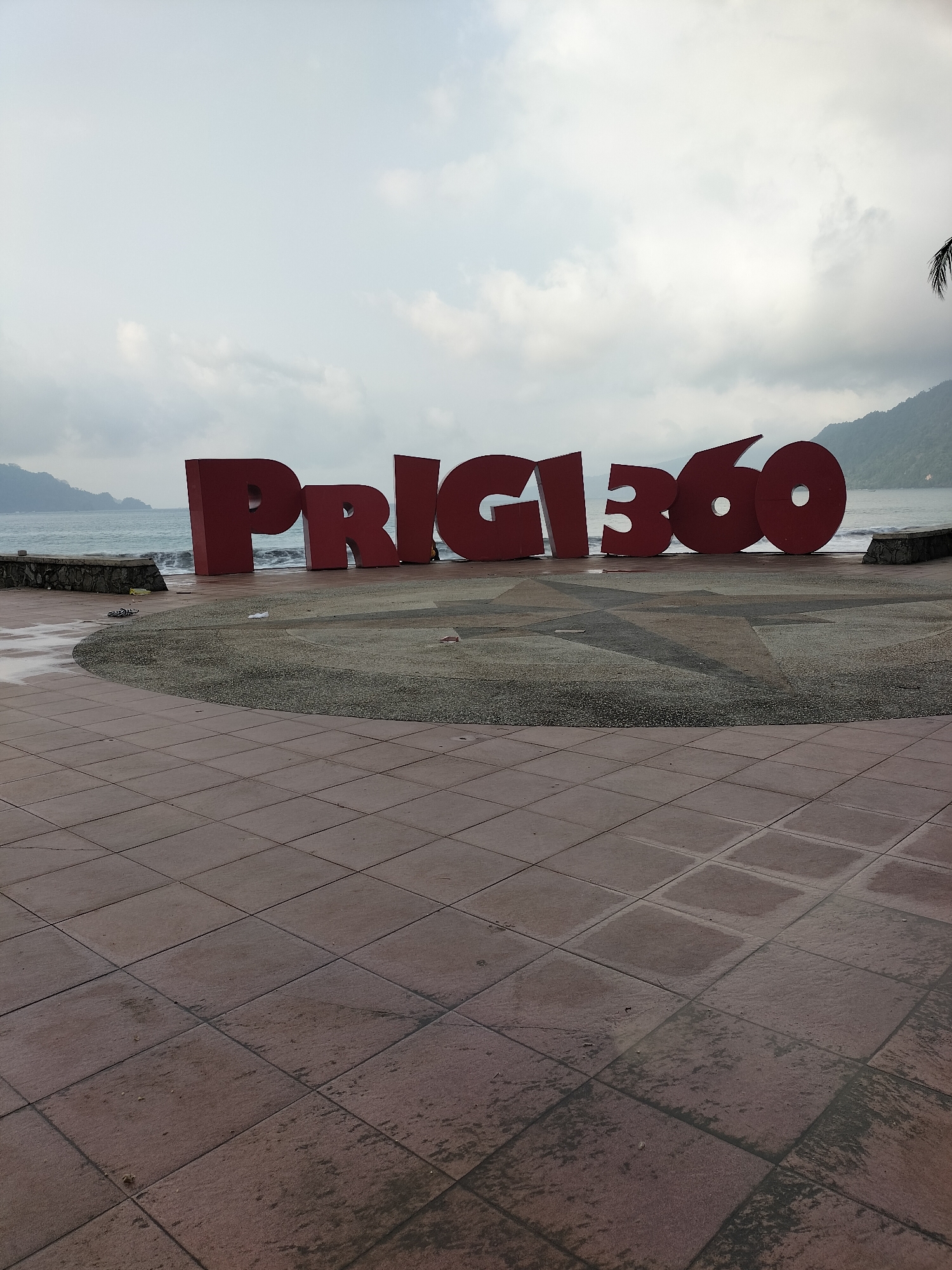 This is a wide-angle, eye-level shot of the "PRIGI360" sign in Tasikmadu, Indonesia, on an overcast day. The sign, composed of large, red, three-dimensional letters, is situated on a paved area with a circular, pebbled section in front of it. Beyond the sign, a calm body of water, likely the ocean, is visible with gentle waves lapping at a stone retaining wall. Mountains covered in dense vegetation form the background on the right side of the image, with a sliver of a palm frond visible at the top right corner. The foreground is dominated by a tiled walkway, with some tiles appearing wet, suggesting recent rain. The sky is heavily clouded, with patches of lighter sky visible. No people are prominently featured in the image, though a small figure in yellow is visible near the right side of the sign. Scattered debris, including what appear to be flip-flops and other small items, are present on the ground near the sign and the retaining wall. The overall impression is a somewhat muted and serene coastal scene, with the bold red sign serving as a focal point.