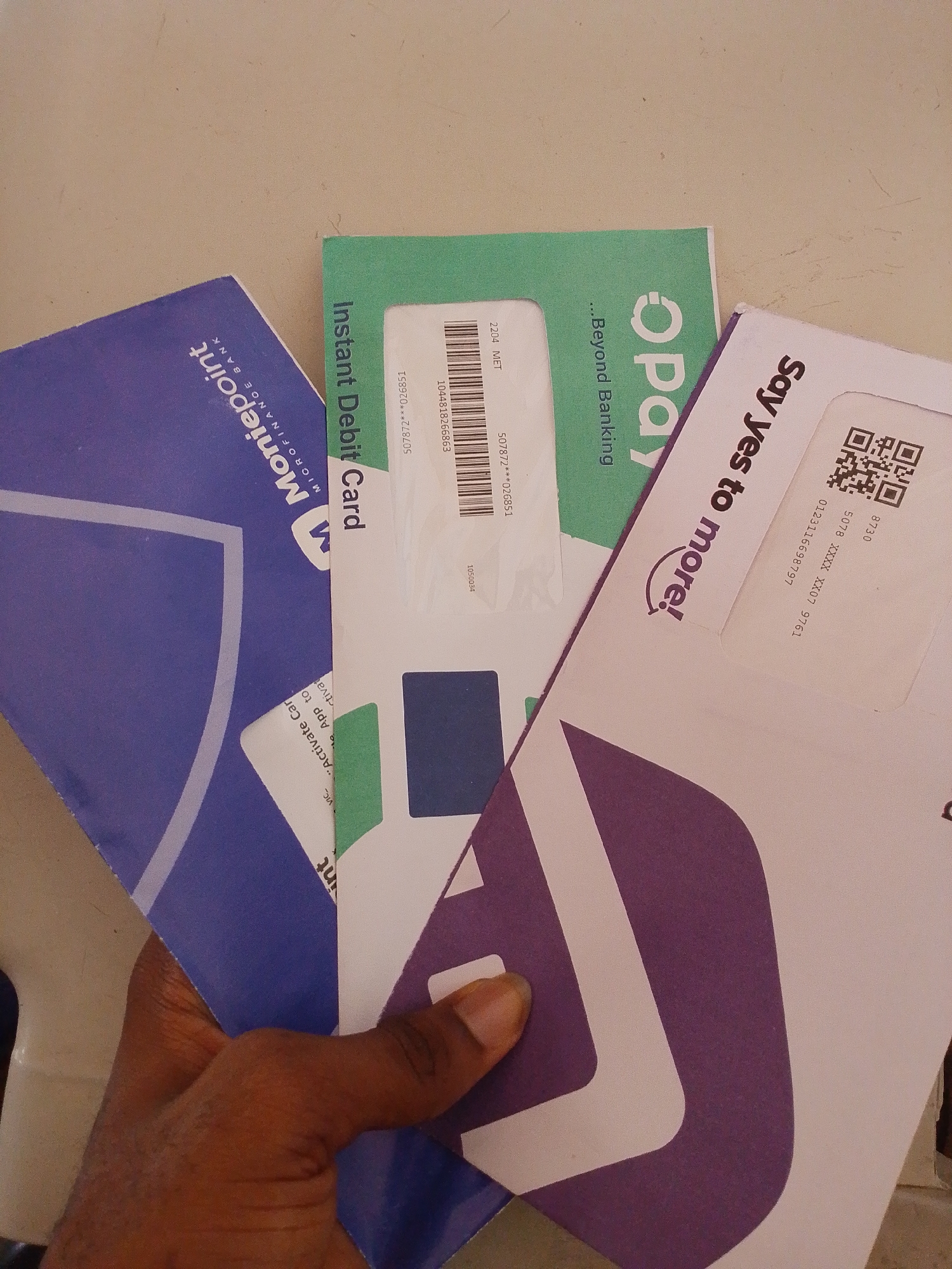 The image shows a hand holding three envelopes, suggesting a financial or banking context. One envelope is green and says "Instant Debit Card" and "...Beyond Banking". Another envelope is purple and says "Moniepoint MICROFINANCE BANK". The third envelope is white with a purple design and the text "Say Yes to more!". The scene is likely indoors, with a neutral background. The lighting is soft and appears to be natural light. The image is taken from a slightly elevated angle, looking down at the hand and envelopes. The envelopes are layered on top of each other, with the green one on top, followed by the purple and white envelopes. The hand is holding the envelopes from the bottom, with the fingers visible. The envelopes appear to be standard letter size. The overall impression is one of official documents being presented or handled.