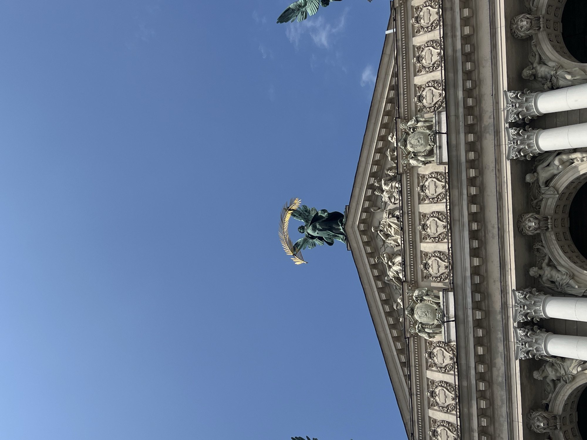 The image shows the top of the Lviv Opera House, also known as the Solomiya Krushelnytska Lviv State Academic Theatre of Opera and Ballet, located in Lviv, Ukraine. A clear blue sky with a few wispy clouds forms the background. At the apex of the building's pediment stands a bronze statue of a winged figure holding a golden laurel wreath, symbolizing victory or fame. To the left and right of this central statue, partially visible, are two other winged figures, also appearing to be statues. The facade of the opera house below the pediment is richly decorated with sculptures of figures, coats of arms, and ornate architectural details, including columns topped with elaborate capitals. The style is Neoclassical or Baroque, characteristic of grand European opera houses.