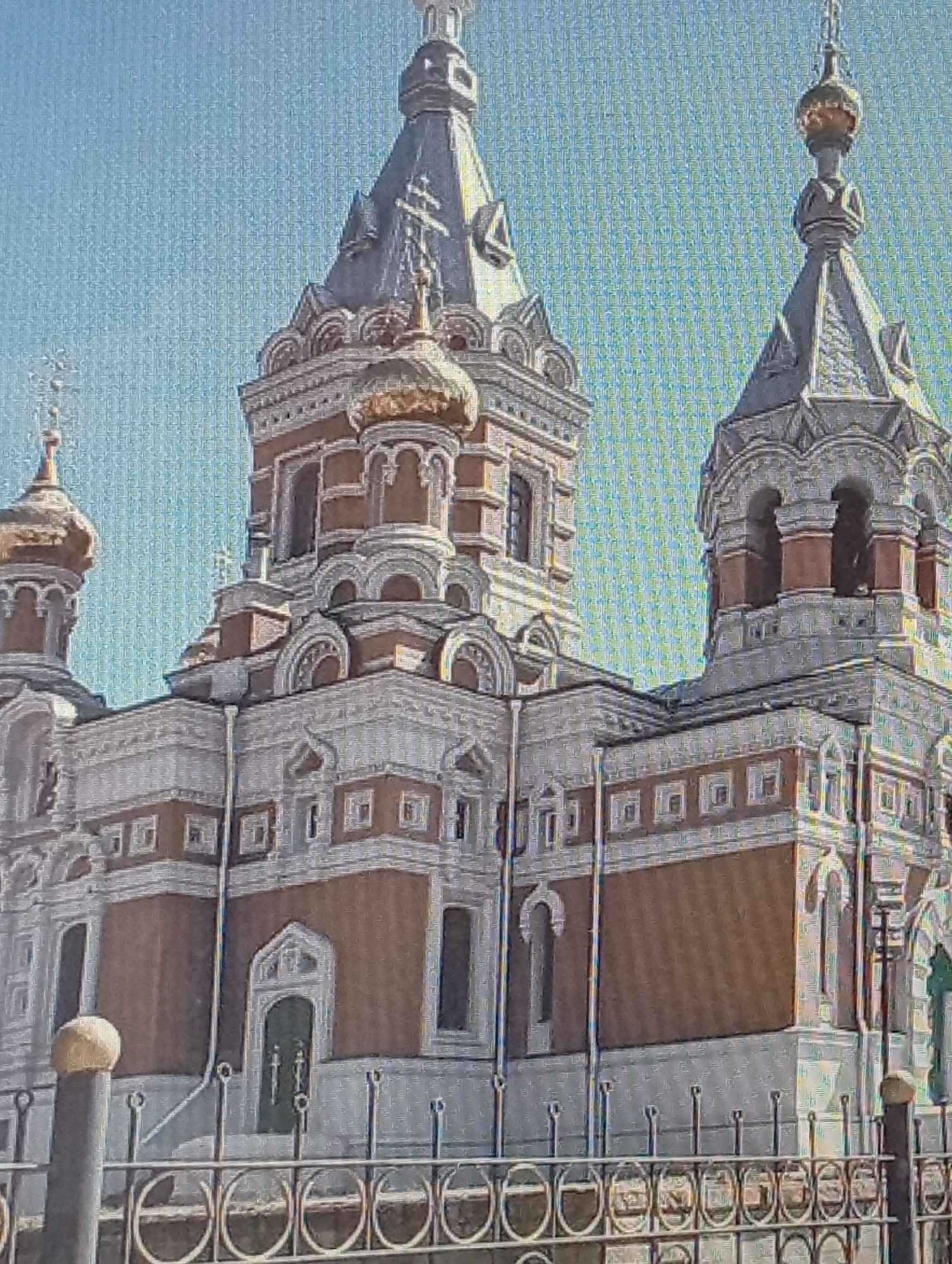 The image shows an exterior view of the Church of the Intercession of the Holy Virgin in the city of Oral, Kazakhstan. The church is a large, ornate building with multiple domes, towers, and intricate architectural details. The main body of the church is clad in brown brick with white decorative elements. It features several arched windows and doorways, as well as decorative friezes and cornices. A prominent golden dome with a cross tops one of the towers. In the foreground, there is a decorative metal fence. The sky is a clear blue. No people are visible in the image.