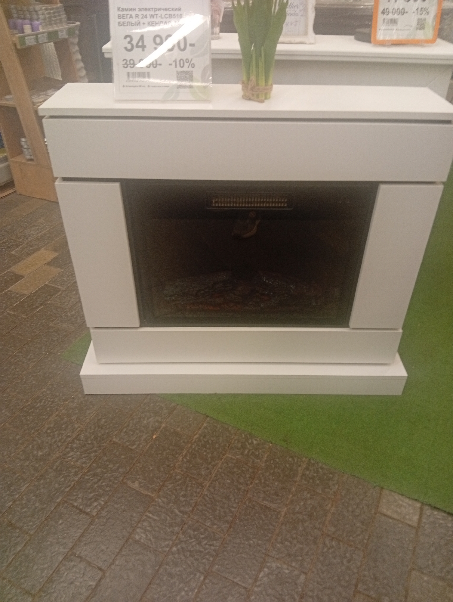 This is a medium shot of a white electric fireplace with a decorative log insert, displayed on a green carpeted floor. The fireplace is the primary subject, and its brand name and model, "Камин электрический ВЕГА R 24 WT-LCB510 БЕЛЫЙ + КЕНДЛ", along with pricing information, are visible on a sign above it. The sign indicates a sale price of 34,990 rubles, a previous price of 39,990 rubles, and a discount of 10%. To the left of the fireplace, a wooden shelving unit with various items is partially visible. The floor in front of the fireplace is tiled. The scene appears to be an indoor retail environment.
