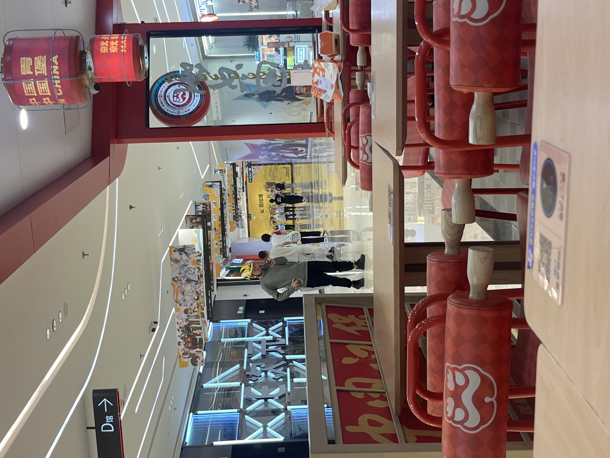 The image depicts a restaurant interior in the Yuzhong District, China. In the foreground, two light-colored tables are visible, one with a pink mat containing a QR code. Red chairs with a distinctive patterned design and a white circular emblem are placed around the tables. In the middle ground, a man wearing a yellow hard hat and a gray long-sleeved shirt is standing, looking at his phone. He is walking through a wide hallway with bright white lighting and a polished floor. Various shops and storefronts line the hallway, with banners and signs visible. One sign, in Chinese characters, indicates "D 馆" with an arrow pointing to the right. Red lanterns with Chinese inscriptions, including "MADE IN CHINA," are suspended from the ceiling. The overall environment appears to be a modern shopping mall or food court.