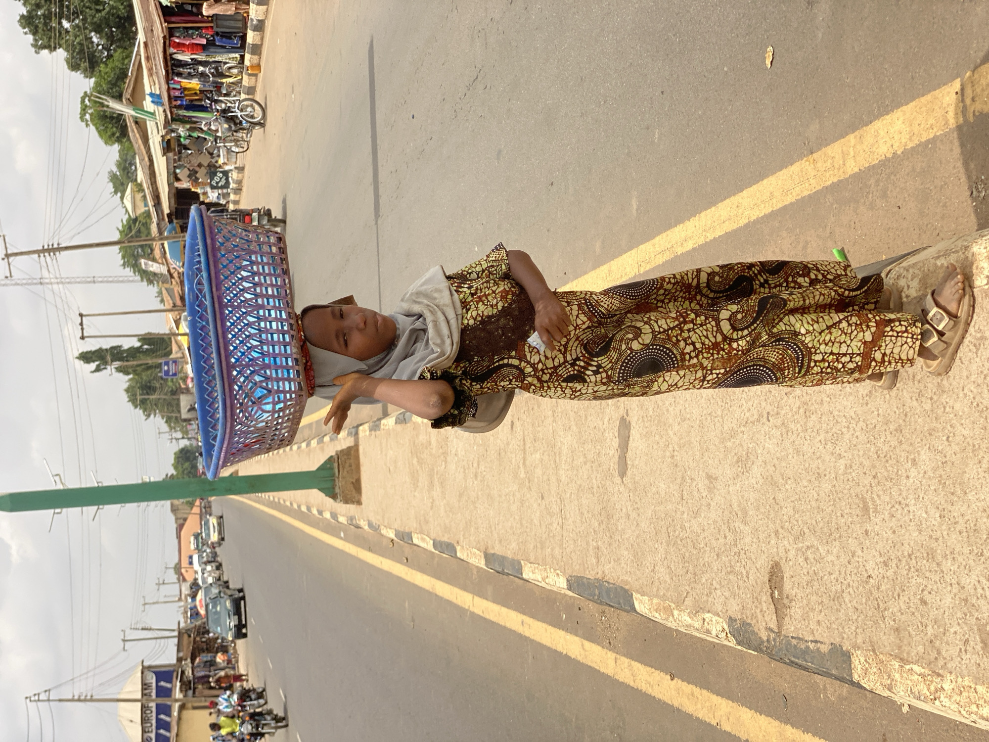 This image captures a young woman or girl standing by the roadside in Kafanchan B, Nigeria, under bright sunlight. She is positioned centrally, facing the camera with a neutral expression. She wears a long, patterned dress or skirt and top in earthy tones of brown, black, and cream, complemented by a light-colored headscarf and sandals. A prominent blue plastic basket is balanced on her head, a common method for carrying goods in the region. She holds a small, white, rectangular object in her left hand.
The setting is a paved street with a clear yellow line running parallel to the curb, which is painted with alternating black and white sections further along. A tall, green metal pole stands to her left. The bright sun creates distinct shadows, suggesting a mid-morning or late afternoon time.
In the background, across the street, a bustling scene unfolds with market stalls or shops displaying various goods. Several motorcycles and a few cars are visible, either parked or in motion. Overhead utility poles and numerous power lines are silhouetted against a pale sky. Some faint text on a sign in the background appears to read "SOS" and "EUROFOAM AMC," possibly indicating local businesses. The image conveys a typical daily scene from a lively Nigerian street.