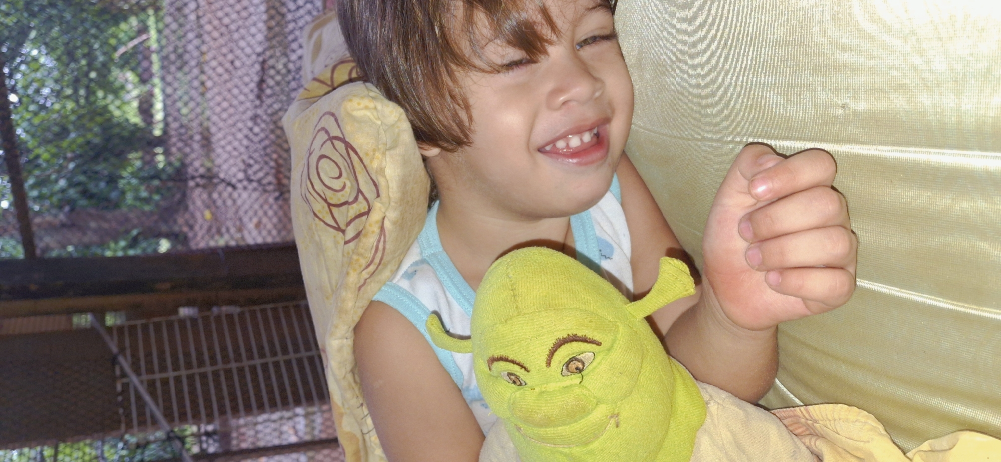 A young child is asleep in bed, covered by a yellow blanket with a floral pattern. The child is lying on their side, facing towards the camera, with their eyes closed and a slight smile on their face. A green stuffed toy, resembling Shrek, is positioned near the child's head. The child is wearing a white t-shirt with a blue trim. The background shows a blurred view of trees and a fence, suggesting an outdoor setting, possibly in a residential area of Vallecito, Venezuela.