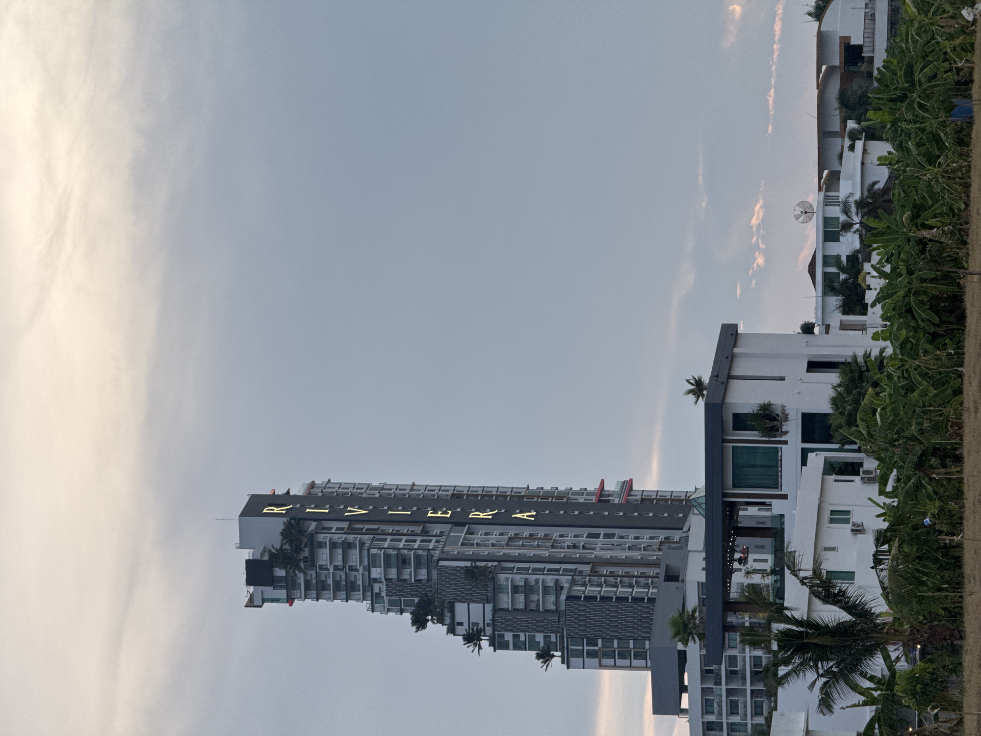 A tall, modern high-rise building, prominently featuring the name "RIVIERA" in illuminated gold letters near its top, dominates the left side of the frame. The building is characterized by numerous balconies and a contemporary architectural style, with grey and white facades. To the right of the skyscraper, several smaller, white, multi-story buildings with flat roofs and dark-framed windows are visible. Some of these adjacent buildings have satellite dishes mounted on their rooftops.
Lush, green tropical vegetation, including palm trees and what appear to be banana plants, is abundant throughout the scene, particularly on the right side, partially obscuring the smaller buildings and framing the view. The sky is partly cloudy, transitioning from a brighter, possibly sun-kissed area on the far left to a softer, uniform light blue-grey towards the right. The soft, diffuse lighting suggests the image was captured during either late afternoon or early morning. The scene depicts a modern urban or resort environment, consistent with Pattaya City, Thailand. No specific activities or individuals are visible.