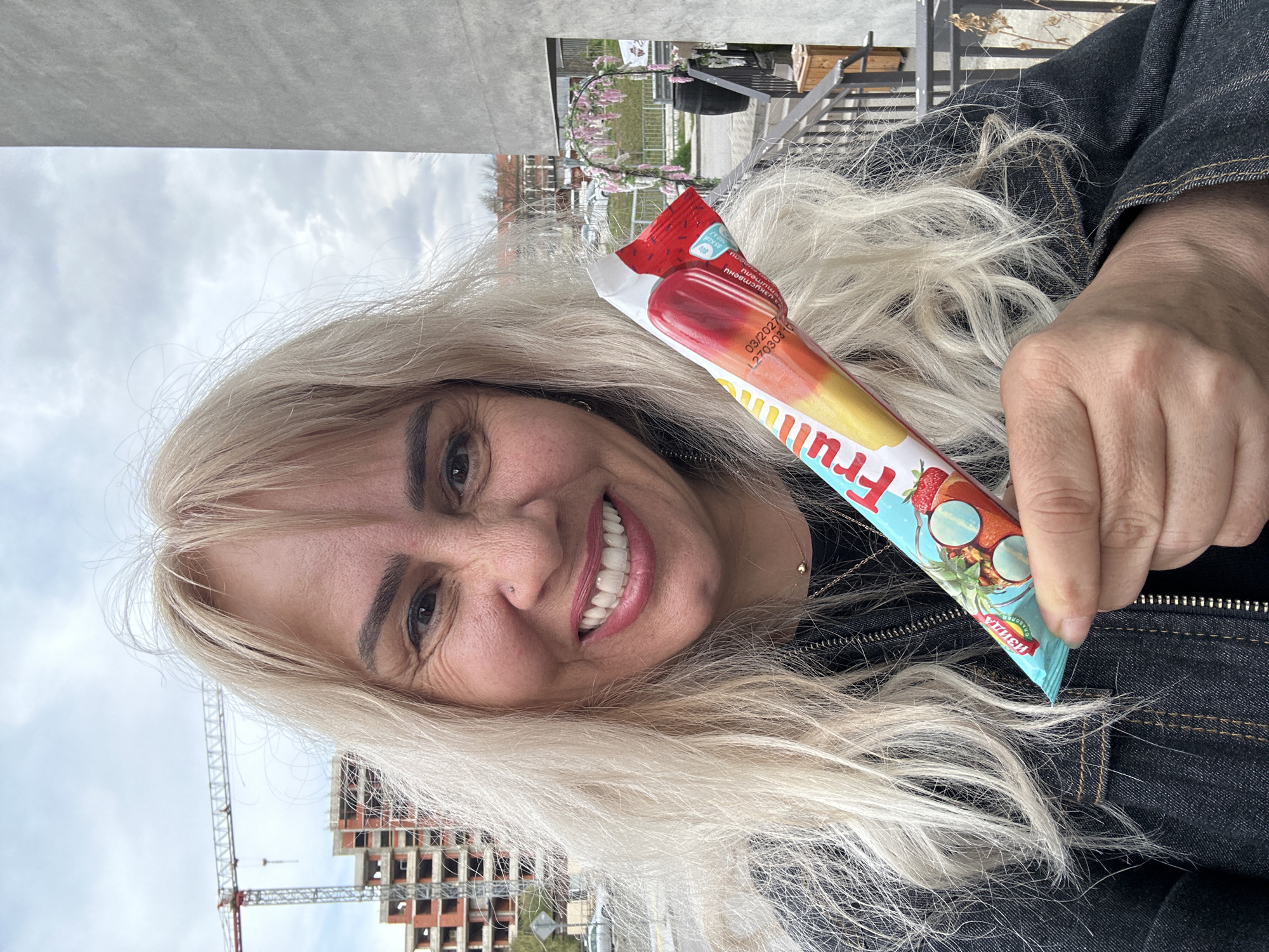 A smiling woman with long, flowing blonde hair is featured in a close-up, selfie-style photograph taken outdoors. She looks directly at the camera with a wide, joyful smile, appearing to be in her 40s or 50s, and is wearing a dark denim jacket. In her right hand, she holds a colorful ice pop. The ice pop wrapper is predominantly red, orange, and blue, featuring illustrations of a strawberry, coconut, and pineapple. The brand name "Fruity" is clearly visible in white letters, along with the expiry date "03/2027" and lot number "L27030310". Partially visible Cyrillic text, including "сладолед" (ice cream), is also on the packaging.
The background shows an urban environment in Sofia, Bulgaria, under an overcast sky. Several buildings are under construction, with visible concrete frameworks and scaffolding. At least two large construction cranes tower over these unfinished structures. In the mid-ground, a decorative archway adorned with pink flowers suggests a landscaped area or public space. The photograph captures a casual daytime moment, with the woman's cheerful expression conveying happiness and relaxation amidst the city's development.