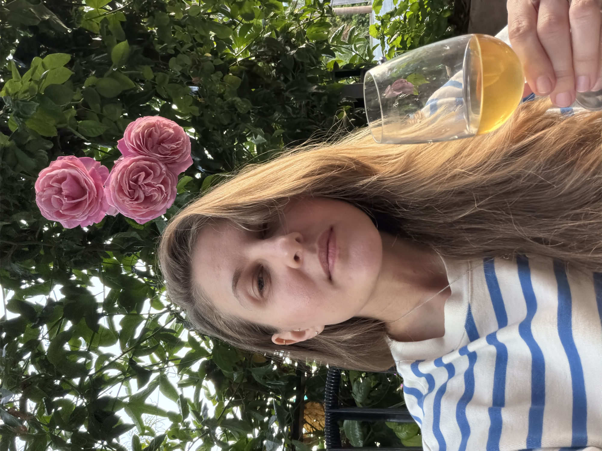 The image depicts a woman holding a glass of liquid, likely wine, with three pink roses visible in the background. She appears to be outdoors, with leafy green foliage forming a backdrop. She is wearing a striped shirt. The exact location within Manhattan Beach, California, cannot be determined from the image.