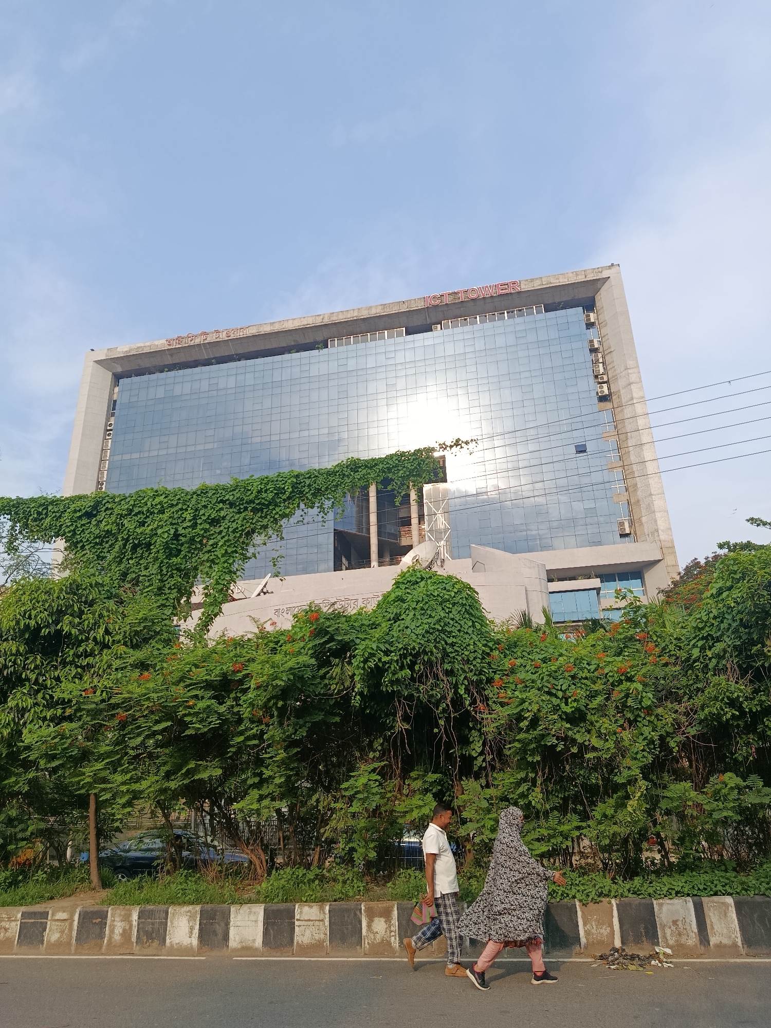 The image depicts a street scene in Dhaka, Bangladesh. In the foreground, a man and a woman are walking along a road with a black and white striped curb. The woman is wearing a floral pattern hijab. Behind them, lush greenery obscures a portion of a large, modern office building with a glass facade reflecting the sky. The building is identified as "ICT TOWER" by signage on the top. Vines are seen growing over the building's exterior. Several utility wires stretch across the sky in the background. A partially hidden car is visible behind the greenery.